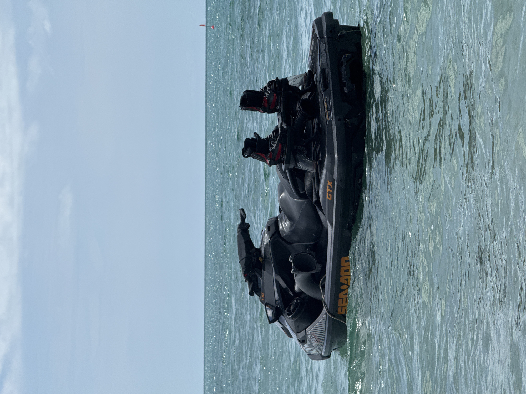 A black Sea-Doo jet ski is floating on the surface of calm, turquoise ocean water under a pale blue sky with scattered clouds. The jet ski is equipped with two black and red wakeboard bindings at the rear. The word "SEN-DOO" is visible in orange lettering along the side of the jet ski, along with the model designation "GTX". The scene appears to be in an open body of water, likely off the coast of Ko Samui District, Thailand, given the clarity of the water and typical aquatic activities in the region. There are no people visible on or around the jet ski. A small red buoy is visible in the distance on the horizon.