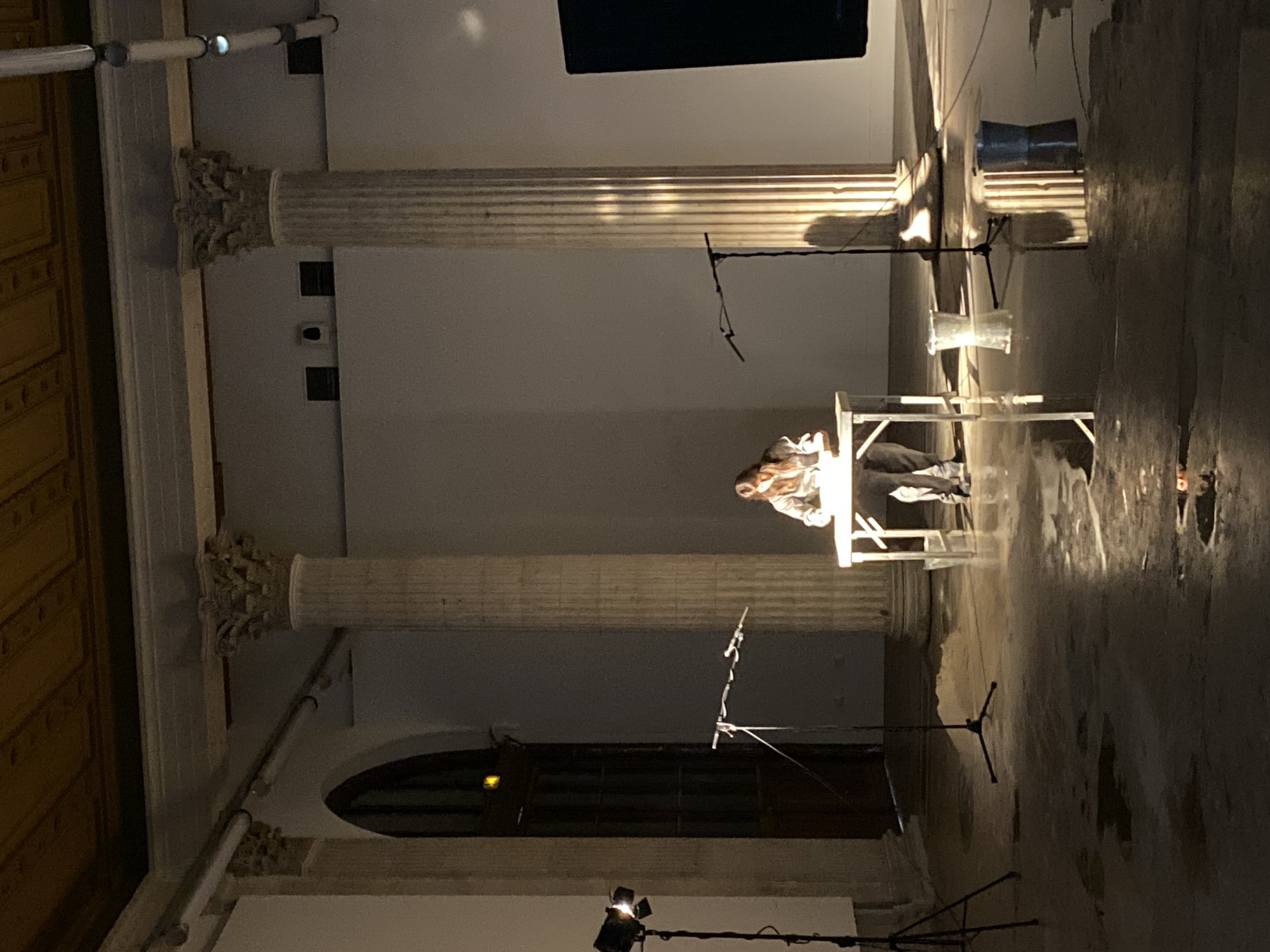 The image captures an indoor scene, likely a performance or art installation, within a grand hall. In the center, a person, possibly a woman with long hair, is seated at a rectangular white table, illuminated by a spotlight on the floor. She appears focused, leaning forward slightly.
The setting is characterized by two large, fluted columns with ornate capitals, flanking the central area. The architectural style suggests a historical or institutional building, consistent with the provided context of Paris, France. To the left, a wall features decorative wooden paneling, while an arched dark doorway or window is visible in the background.
Numerous objects related to a performance or technical setup are present: multiple microphone stands are positioned around the space, along with lighting equipment including the floor spotlight. Wires and cables run across the floor. A dark, cylindrical object, possibly a speaker or container, is seen near one of the columns, and a clear, bucket-like container hangs from a stand. The floor has reflective patches, suggesting either a wet surface or a deliberate reflective element as part of the installation.
The lighting is dramatic, with a strong spotlight creating deep shadows and highlighting the person and table, suggesting it is evening or night, or a purposefully dimmed environment. No visible text is present in the image.
