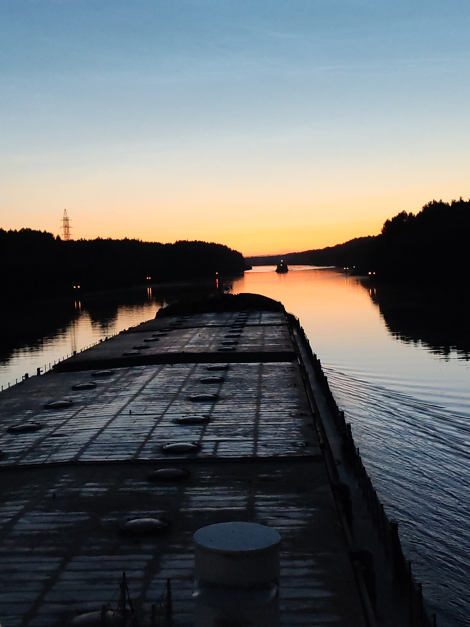 The image shows a wide, calm body of water, likely a river or canal, bordered by dark, silhouetted trees on either side. The sky above is a gradient of soft blues and oranges, indicating either sunrise or sunset. In the foreground, the deck of a barge or ship is visible, extending into the frame from the bottom left. The deck is flat and appears to be made of metal, with a series of circular hatches or covers. The water is dark and reflects the colors of the sky, with gentle ripples suggesting movement. In the distance, two other vessels can be seen on the water, their lights dimly visible. One is a smaller boat or tug, and the other appears to be another barge. A tall, lattice-like structure, possibly a power line pylon, is also visible against the sky on the left bank, in the distance. The overall scene evokes a sense of tranquility and the vastness of nature. The specific location in the city of Aleksandrovskoe, Russia, is not evident from visual cues within the image itself.
