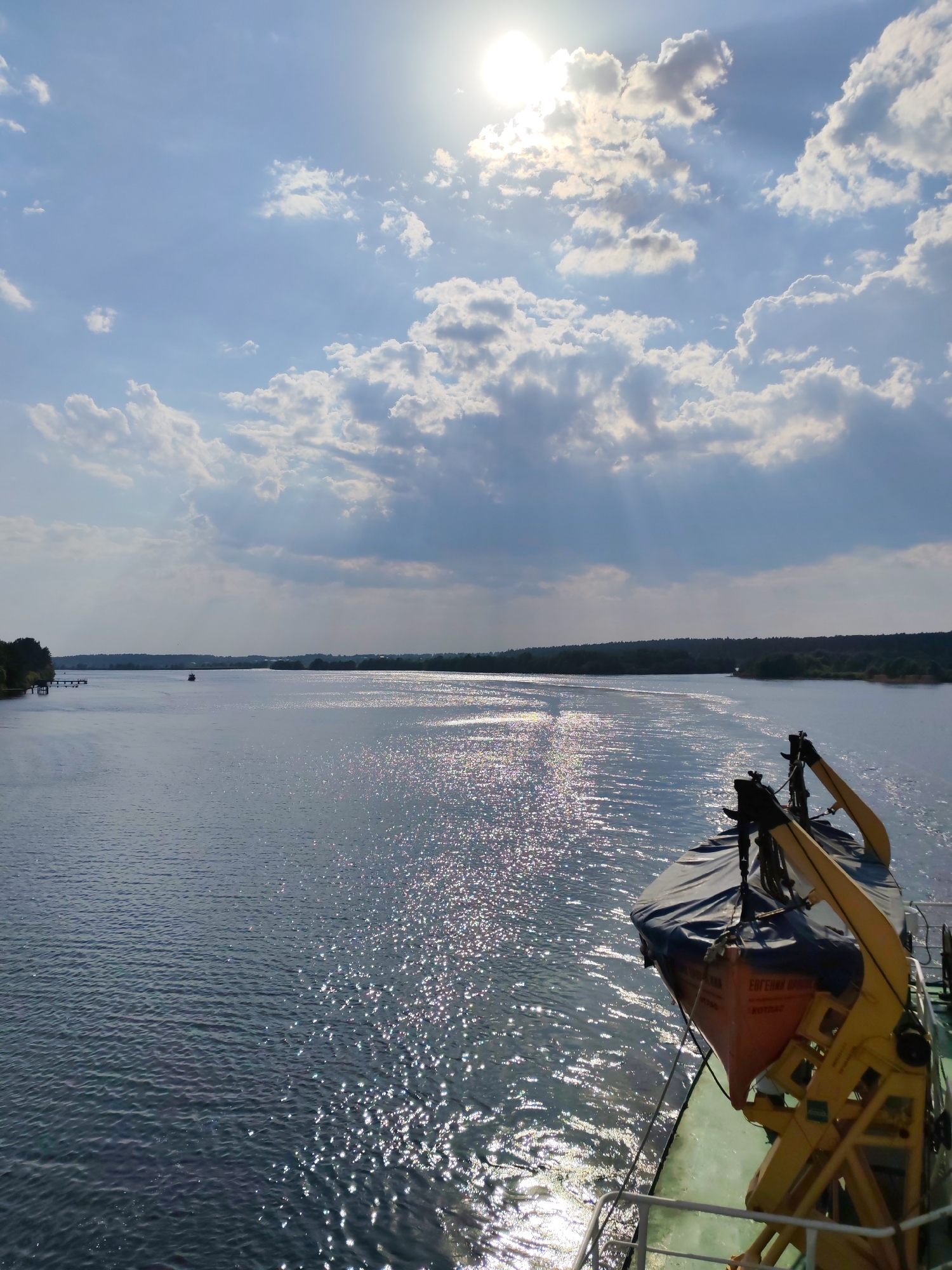 The image captures a scene on the Volga River near Kimry, Russia. The view is from a boat or ship, showing a lifeboat hanging on a yellow davit system on the right side. The Volga River extends into the distance, its surface shimmering with reflected sunlight. The river banks are lined with trees and greenery. The sky is a mix of blue and white clouds, with the sun high above, partially obscured by the clouds. A small boat can be seen on the river in the distance.