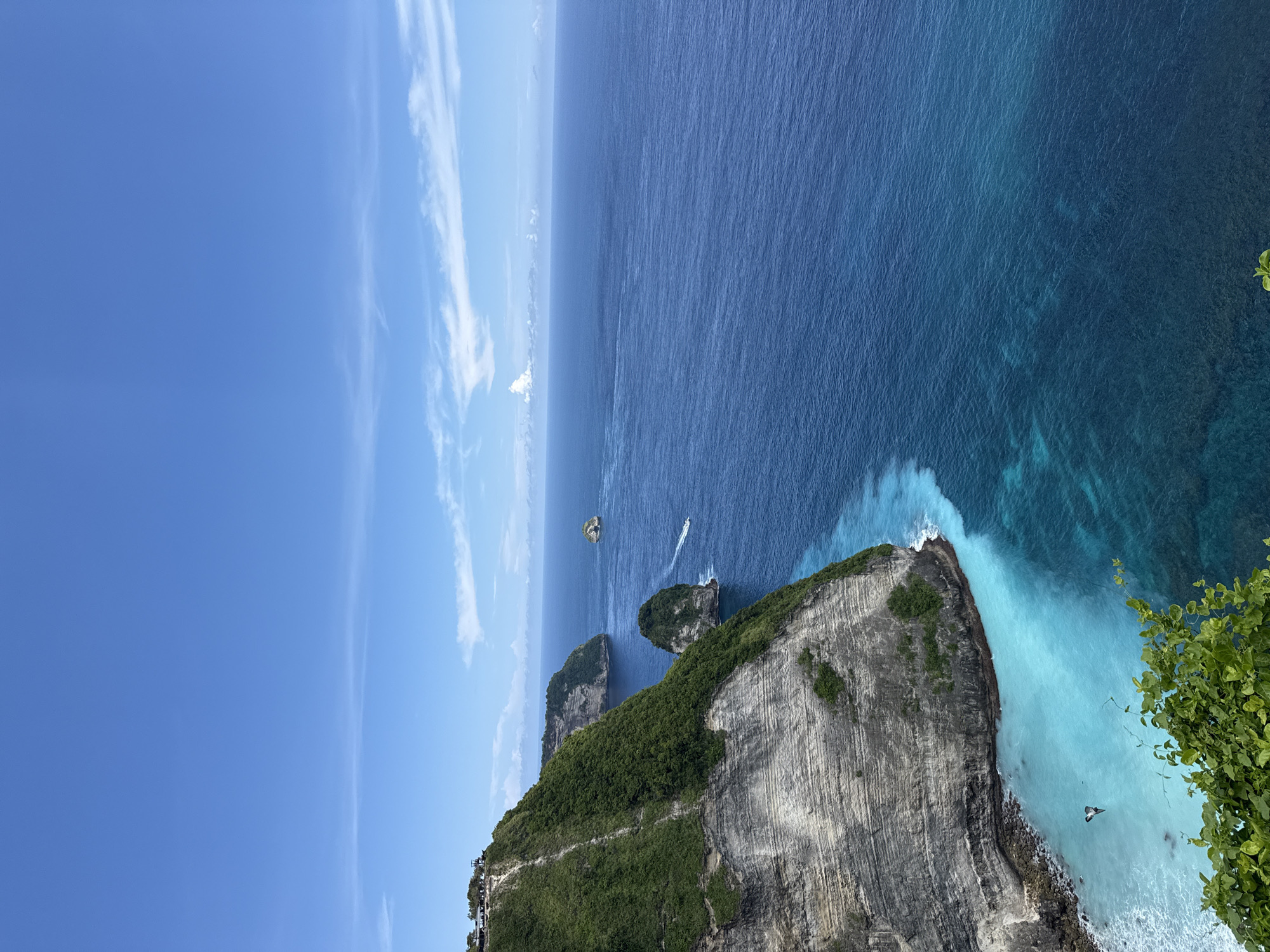 The image shows an ocean view from the cliffside of Nusa Penida, an island southeast of Bali, Indonesia, in the village of Bunga Mekar. The sky is blue with streaks of cirrus clouds. The ocean is a gradient of dark blue in the distance to turquoise near the shore. Several small, rocky islands rise from the sea. A small boat is visible on the water. A high cliff face covered with green vegetation occupies the foreground. A narrow trail is visible on the cliffside. The bottom right corner of the frame contains some green leaves.