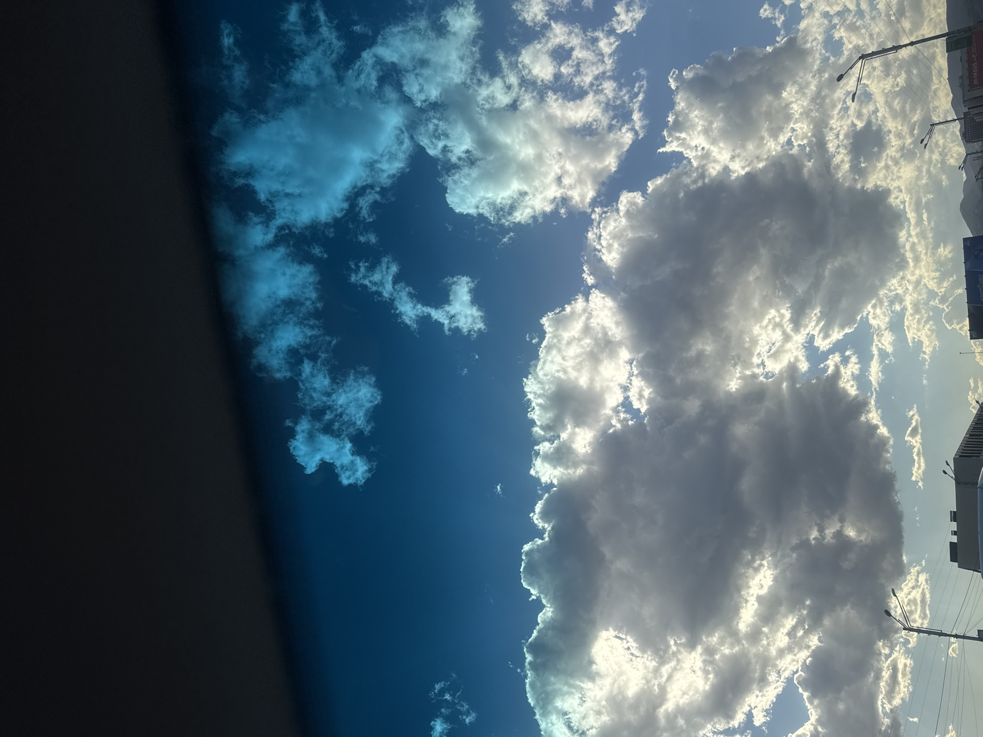 The image depicts a view of the sky above Tbilisi, Georgia. Predominantly blue sky with scattered, fluffy clouds. The clouds vary in color from light gray to bright white. The sun is obscured by the clouds, but its light filters through. Tall, narrow buildings and a street lamp can be seen on the right side of the image, and a dark object that might be part of a building or vehicle is present on the left side.