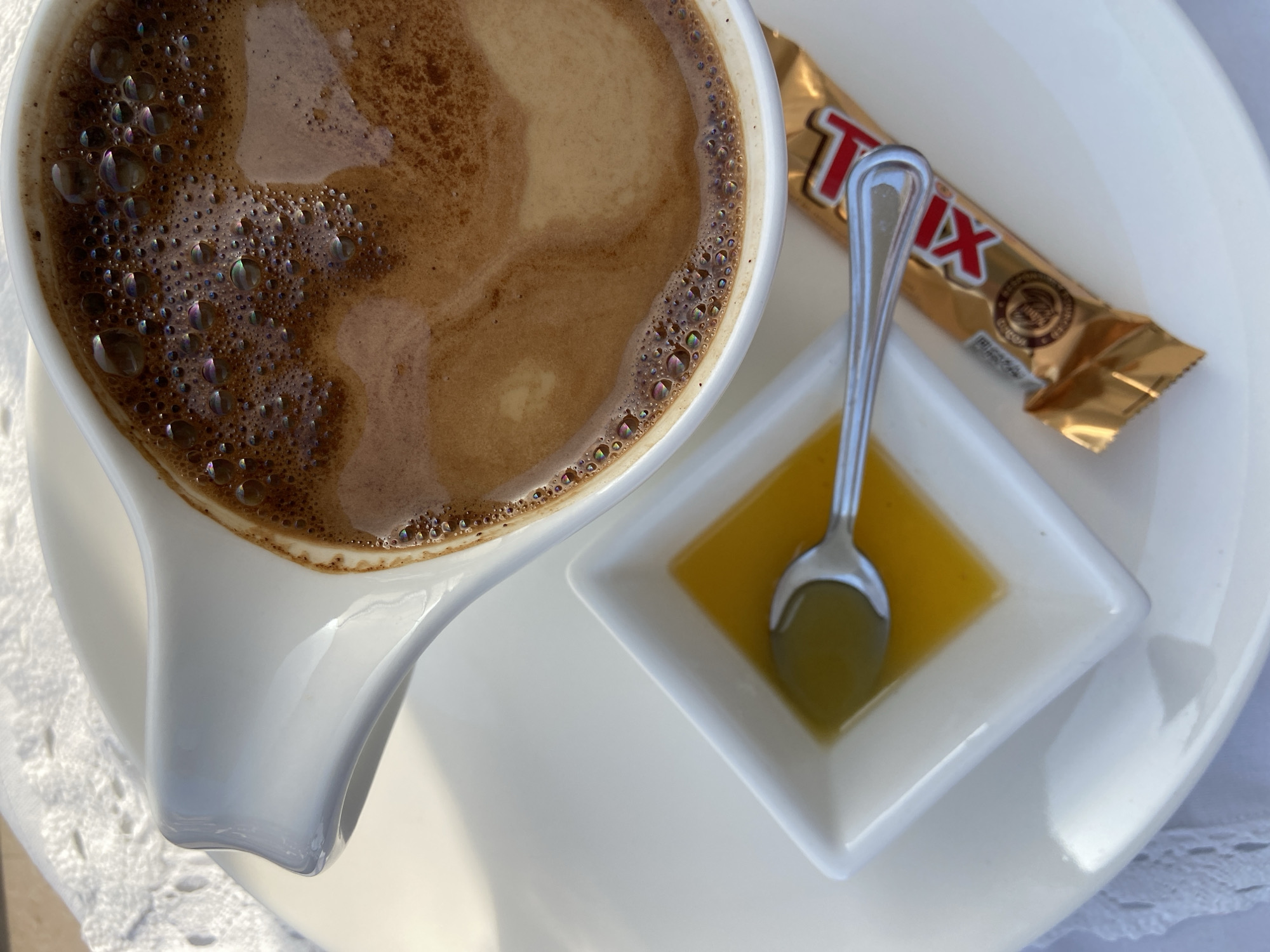 A top-down view captures a coffee cup filled with coffee, accompanied by a small square dish containing honey and a spoon, and a Trix chocolate bar, all placed on a white plate. The coffee has a foamy surface with iridescent bubbles. The honey is yellow-gold, and the Trix bar has a gold wrapper with red lettering. The cup is white with a distinctive spout. The scene is set on a white tablecloth. The location is Budva, Montenegro.