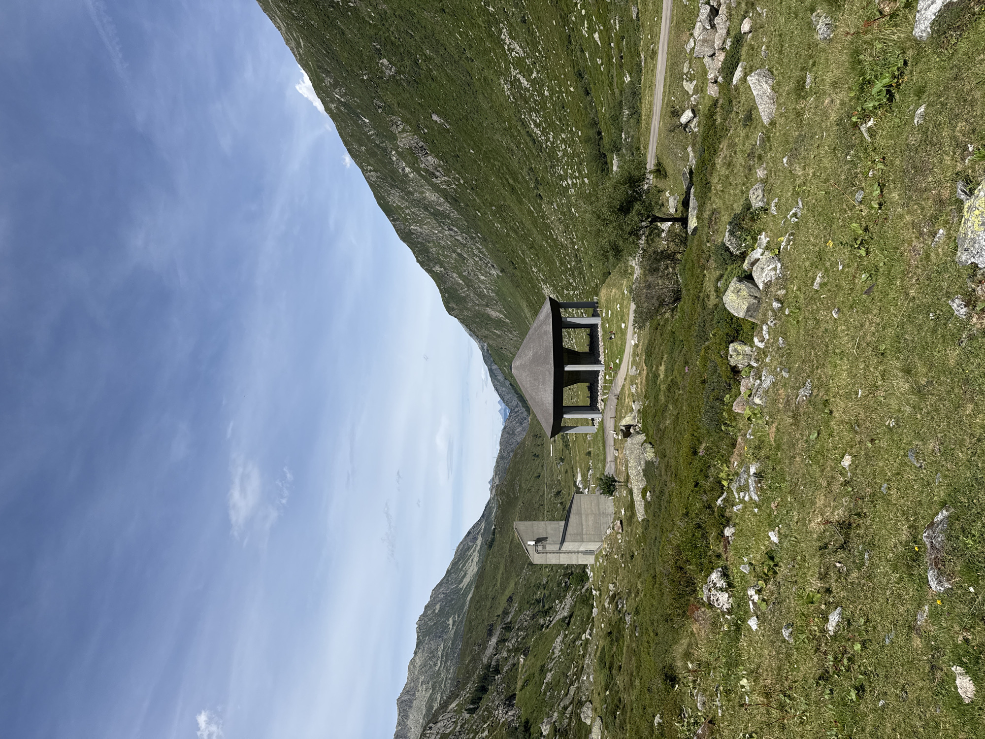 The image depicts a scenic mountain landscape in Hospental, Switzerland. In the foreground, a grassy slope is dotted with large rocks and sparse vegetation. A paved road runs through the middle ground, bordered by more rocks and low-lying green plants. Two prominent structures are visible: on the left, a modern, angular concrete building with a prominent vertical antenna, and on the right, a circular, open-sided pavilion with a dark, conical roof supported by pillars. Both structures are situated in a valley surrounded by steep, verdant mountains. The sky above is a bright blue, with scattered wispy clouds. The overall scene is one of natural beauty with architectural elements integrated into the environment.