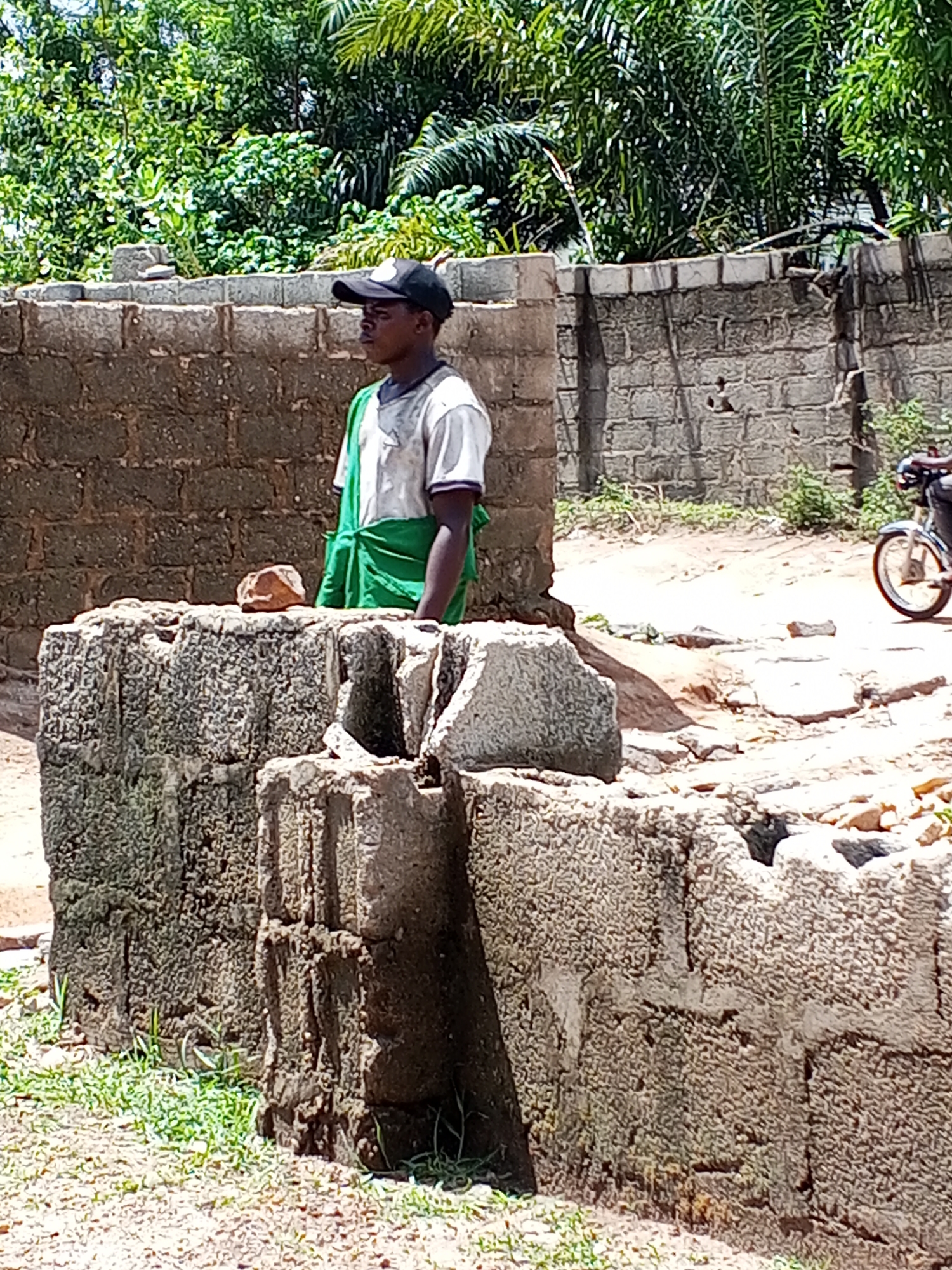 Adonkolo, Nigeria: A young man wearing a black baseball cap and a white t-shirt, with a green cloth draped over one shoulder, stands behind a partially constructed stone structure. A brick wall is visible behind him, and a motorcycle is partially visible on a dirt road in the background. Trees and vegetation are also visible.