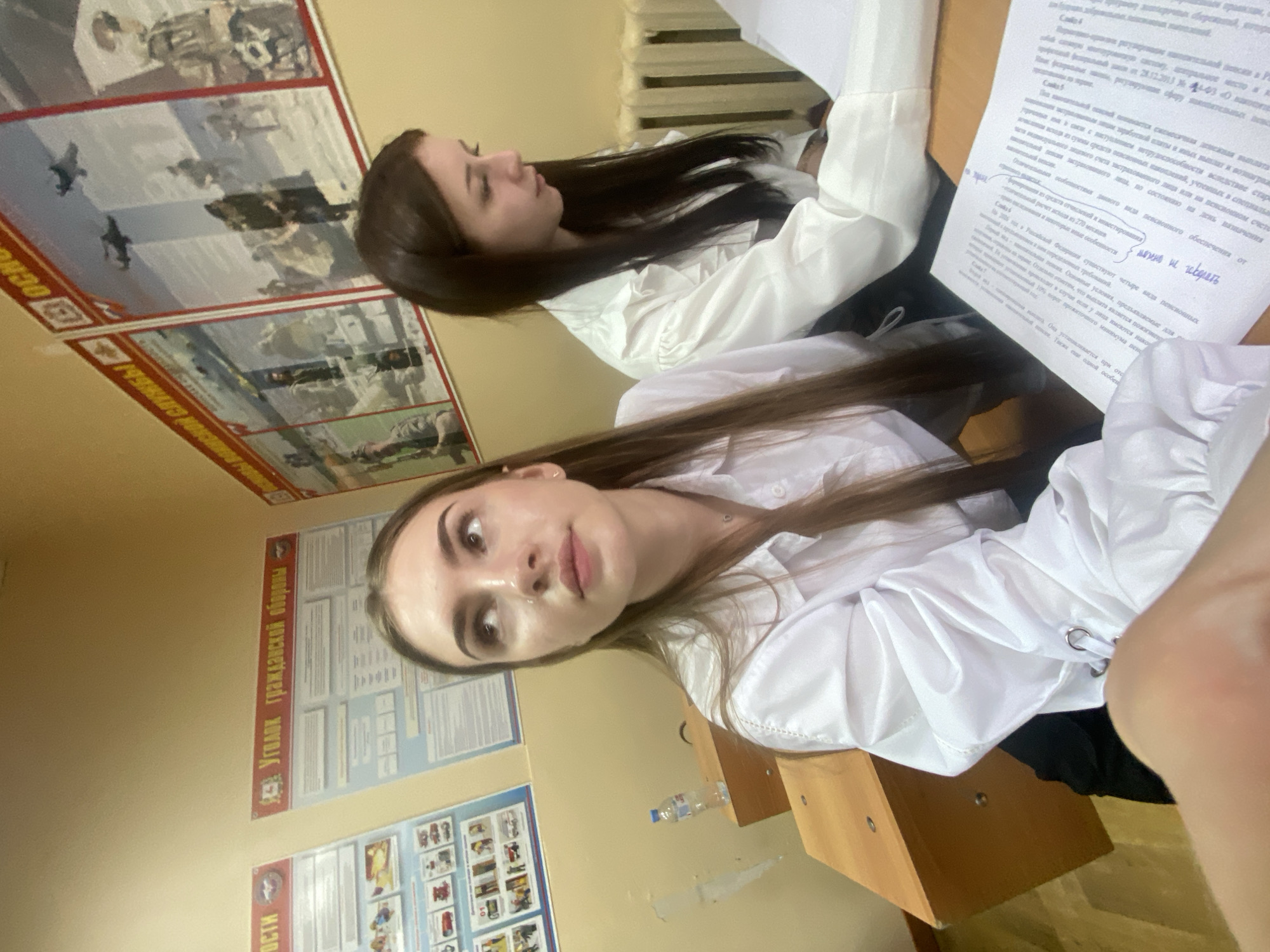The image depicts two young women, likely students, seated at desks in a classroom. The woman on the left is in the foreground, looking upwards and to the right, with her head tilted. She has long, light brown hair and is wearing a white collared shirt. The woman on the right is in the background, looking down at papers on her desk. She has long, dark brown hair and is also wearing a white collared shirt.
The classroom environment is suggested by the presence of desks and educational posters on the wall. One poster in the background appears to have a title that translates to "Corner of Civil Defense." Another poster to the right features images of military aircraft and personnel, with Russian text. A water bottle is visible on the desk to the left. The overall lighting suggests an indoor setting during the day. The provided contextual information states the location is Dyakovo, Russia.