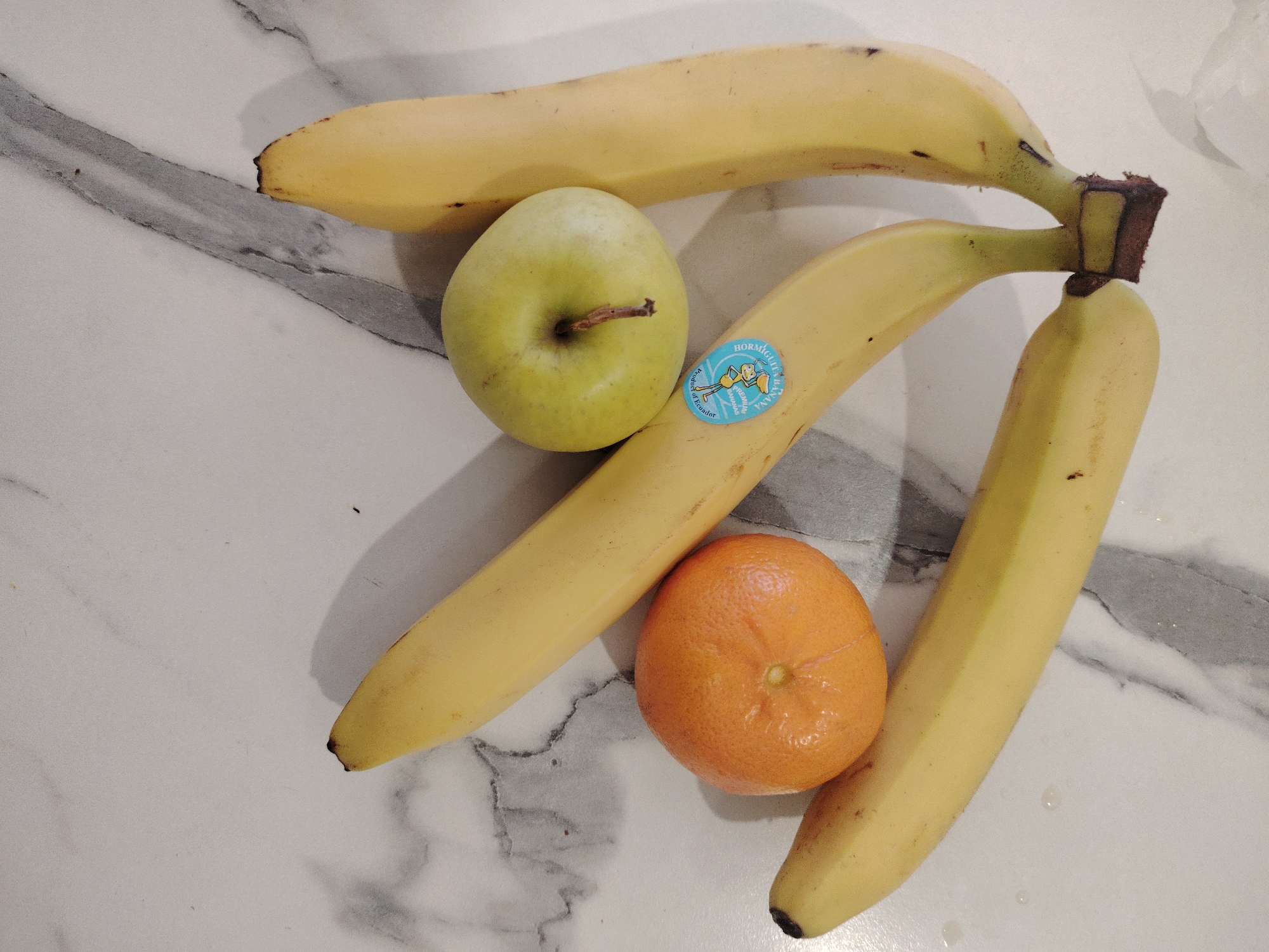 The image presents a still life arrangement of fruits on a white marble countertop. Three bananas are clustered together, their stems connected. An apple, light green with a visible stem, sits near the center of the composition. An orange is positioned near the lower center, displaying a textured skin. A sticker is visible on one of the bananas. The background is the white marble surface with grey marbling. There is no indication of the city of Sarapul, Russia.