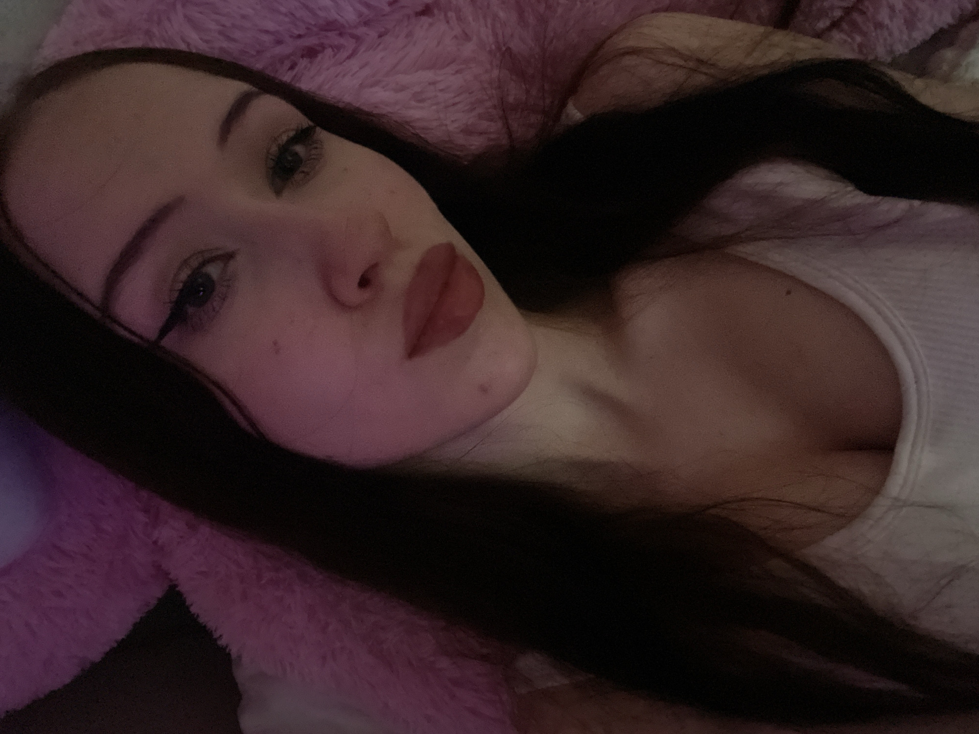 A young woman with dark hair is lying down, with her head and upper torso visible. She is wearing a white tank top and has makeup on, including winged eyeliner and lipstick. Her skin appears pale and is illuminated by soft pink light. She is positioned against a plush pink surface, possibly a blanket or stuffed animal, which is slightly out of focus. The background is dark and indistinct. The image does not provide any specific information about the location in the city of Votkinsk, Russia.