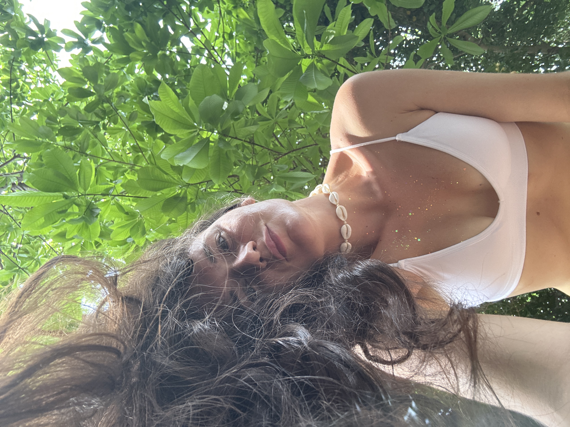 A young woman with long, dark, wavy hair is pictured from a low angle outdoors, with green foliage overhead. She is wearing a white bikini top and a seashell necklace. Her hair is thrown back, obscuring part of her face, and glitter is visible on her chest and shoulders. The background consists of lush green leaves and branches, suggesting a natural, possibly tropical, setting. The lighting indicates it is daytime, with sunlight filtering through the leaves. The overall impression is one of relaxed, outdoor leisure.