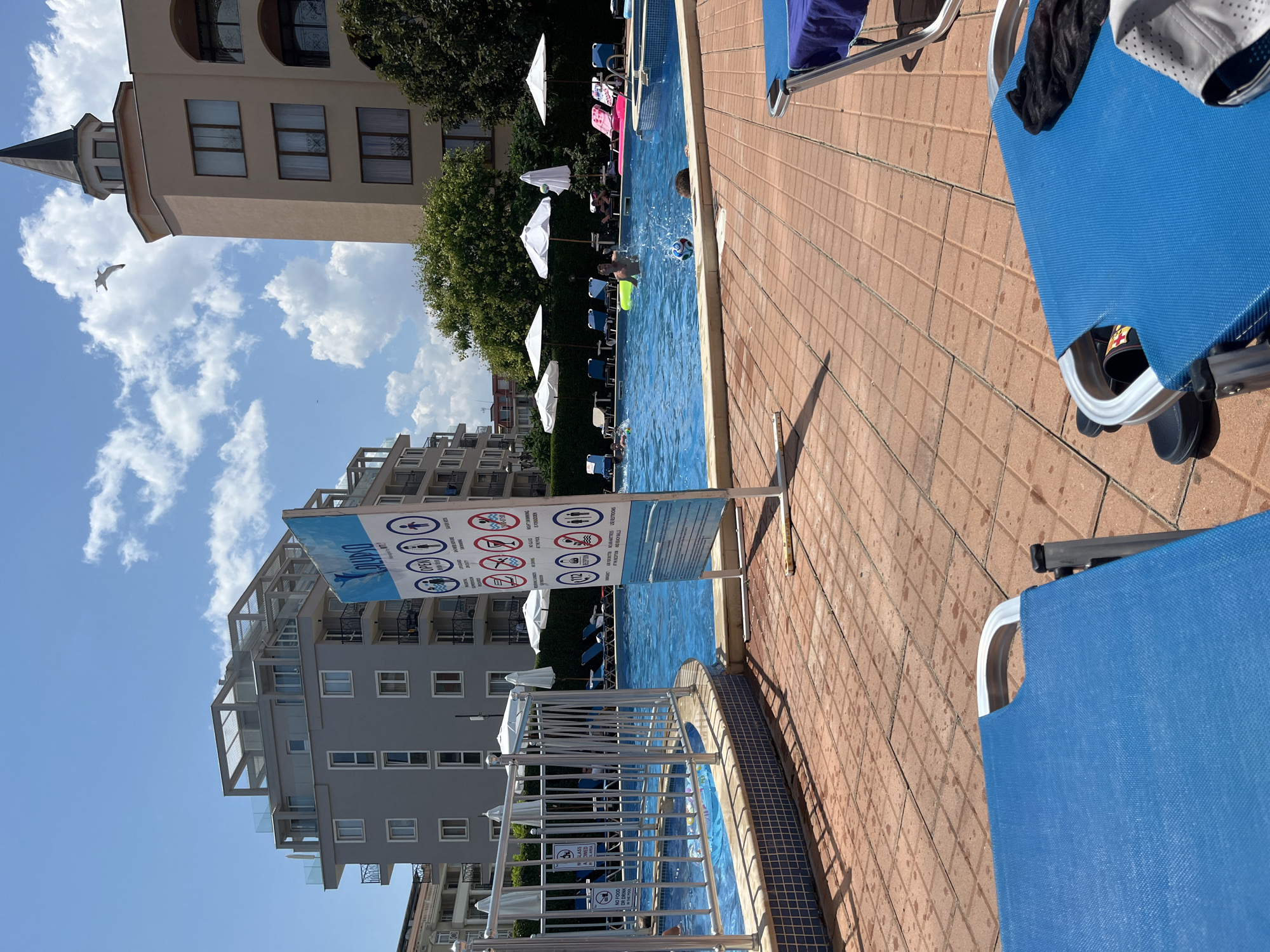 The image depicts an outdoor swimming pool area, likely at a hotel or resort, under a bright, partly cloudy sky. In the foreground, two blue lounge chairs are visible, one in sharp focus and the other slightly blurred. The pool itself is filled with clear blue water, and several people are swimming and playing. One person is in the water, seemingly playing with a ball, while others are visible further back. Around the pool, there are white umbrellas providing shade and lounge chairs for sunbathers. A metal fence encloses a portion of the pool area.
A prominent sign with various icons and text stands near the pool, displaying rules and information. Buildings, typical of a coastal resort town, are visible in the background. These include a modern gray building with balconies and an older, tan building with a distinctive tower and arched windows. Trees and other vegetation surround the pool area. The ground around the pool is paved with reddish-brown tiles. A seagull is captured in flight against the sky, adding to the vacation atmosphere. The overall scene suggests a sunny day at a recreational facility in Nesebar, Bulgaria, a popular Black Sea resort town known for its beaches and historical sites.