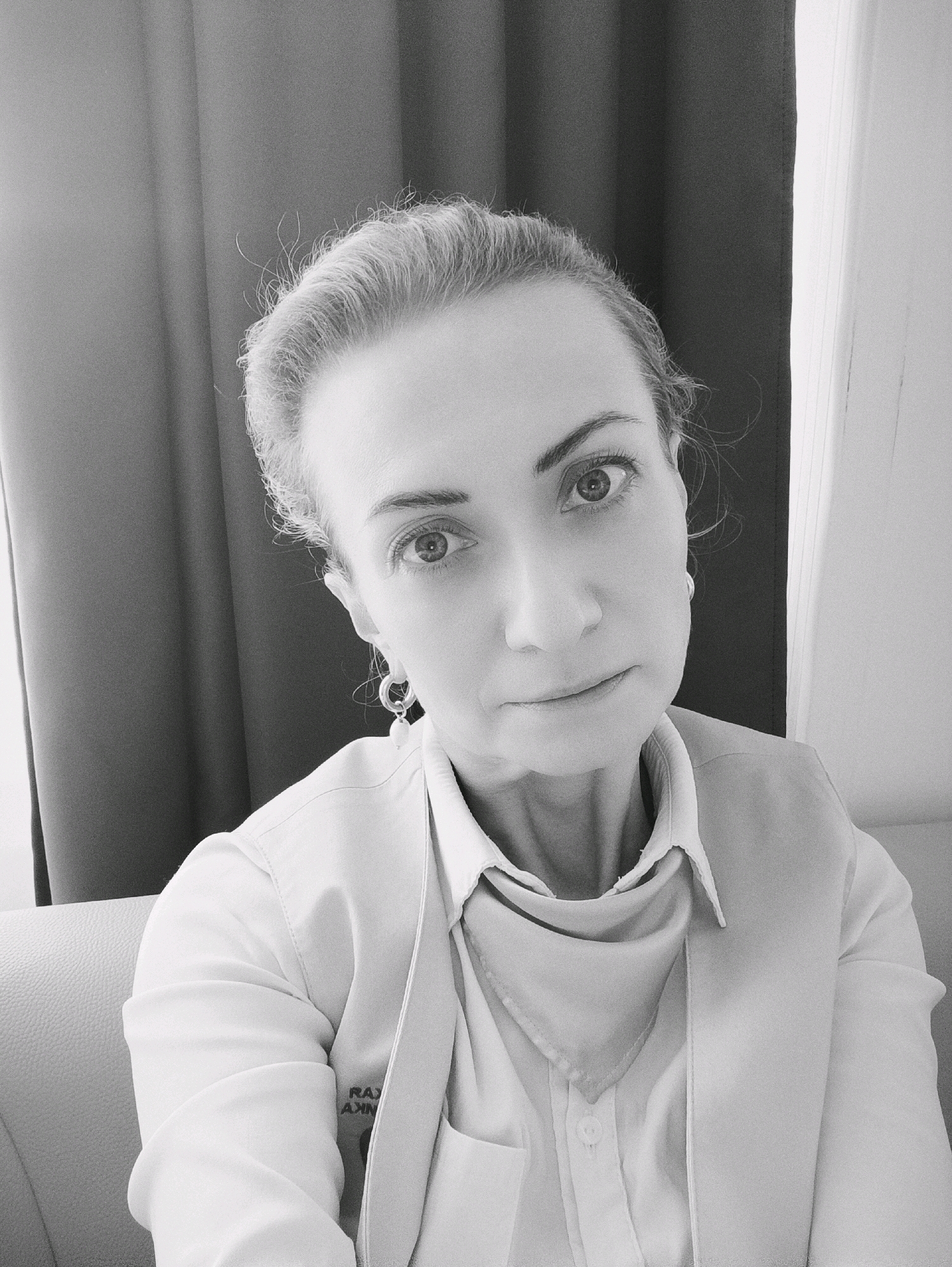 A black and white, medium shot, waist-up, selfie of a woman sitting on a sofa. The woman is facing forward with her head tilted slightly to her right. She has blonde hair pulled back and a small scarf around her neck. She is wearing a light-colored shirt and vest. The background consists of a dark curtain to the left and a light-colored wall to the right. The image appears to have been taken indoors, possibly in an office or hotel room. There is no indication of the specific location within Moscow, Russia.