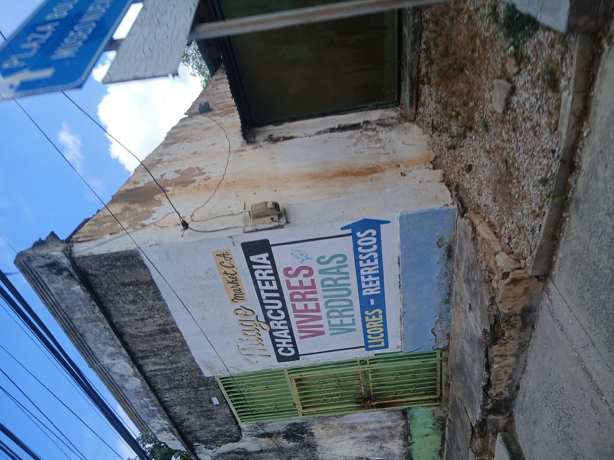 The image shows the facade of a commercial establishment in La Asuncion, Venezuela. The business is named "Tiago's Market C.A." and offers "Charcuteria, Viveres, Verduras, Licores - Refrescos" (Deli, Groceries, Vegetables, Liquor - Soft Drinks). The building appears old and weathered, with peeling paint and crumbling edges. A green, barred gate covers a doorway. Above the store is a window and along the side is a wall of gravel. In the distance, a blue road sign marked "PLAZA BOLIVAR" is visible. Powerlines run overhead.