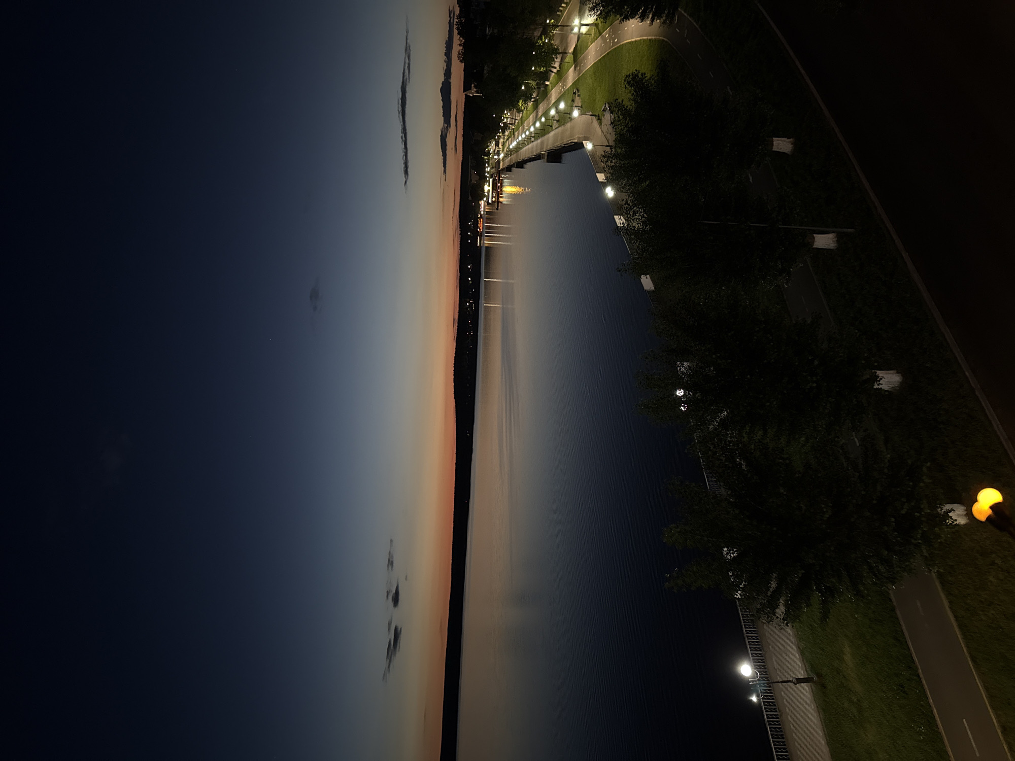The image captures a panoramic view of a waterfront scene at dusk or early night in Votkinsk, Russia. A wide body of water, likely a river or lake, dominates the left side of the frame, its surface reflecting the fading light of the sky and the artificial illuminations from the shore. The sky above transitions from a pale, light orange and blue horizon line to a deep, dark navy blue and black overhead, suggesting the last remnants of sunset giving way to night. Wispy clouds are visible in the lighter part of the sky.
On the right side of the image, a well-lit promenade or park area runs parallel to the water. A long line of streetlights with warm, glowing bulbs extends into the distance, casting bright light onto the paved pathway and grassy areas. Lush trees, appearing dark against the artificial lights, line the promenade. Beyond the trees and pathway, a darker area suggests a further extension of the land or more distant structures. On the far bank of the water, distant lights indicate a settlement or infrastructure. No people are visible in the scene, which appears quiet and tranquil. There is no discernible text. The overall atmosphere is peaceful, highlighting the contrast between the natural twilight and the artificial glow of the city's lights.