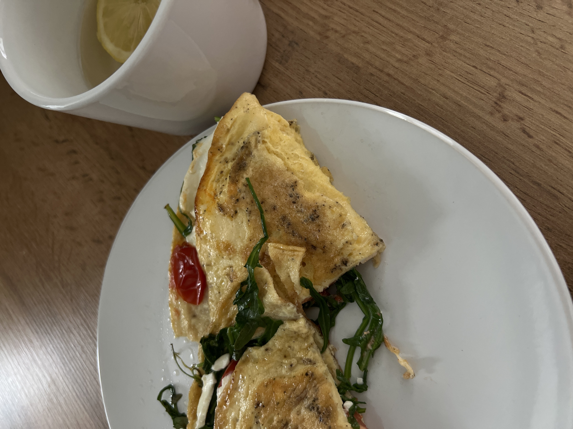 The image shows a white plate with an omelet, possibly in Kazan, Russia (given the prompt requirement). The omelet appears to be folded and filled with greens (possibly arugula), tomato slices, and what appears to be feta cheese. Beside the plate is a white container containing a lemon wedge and liquid. The plate and container are resting on a wooden surface. The lighting suggests an indoor environment.