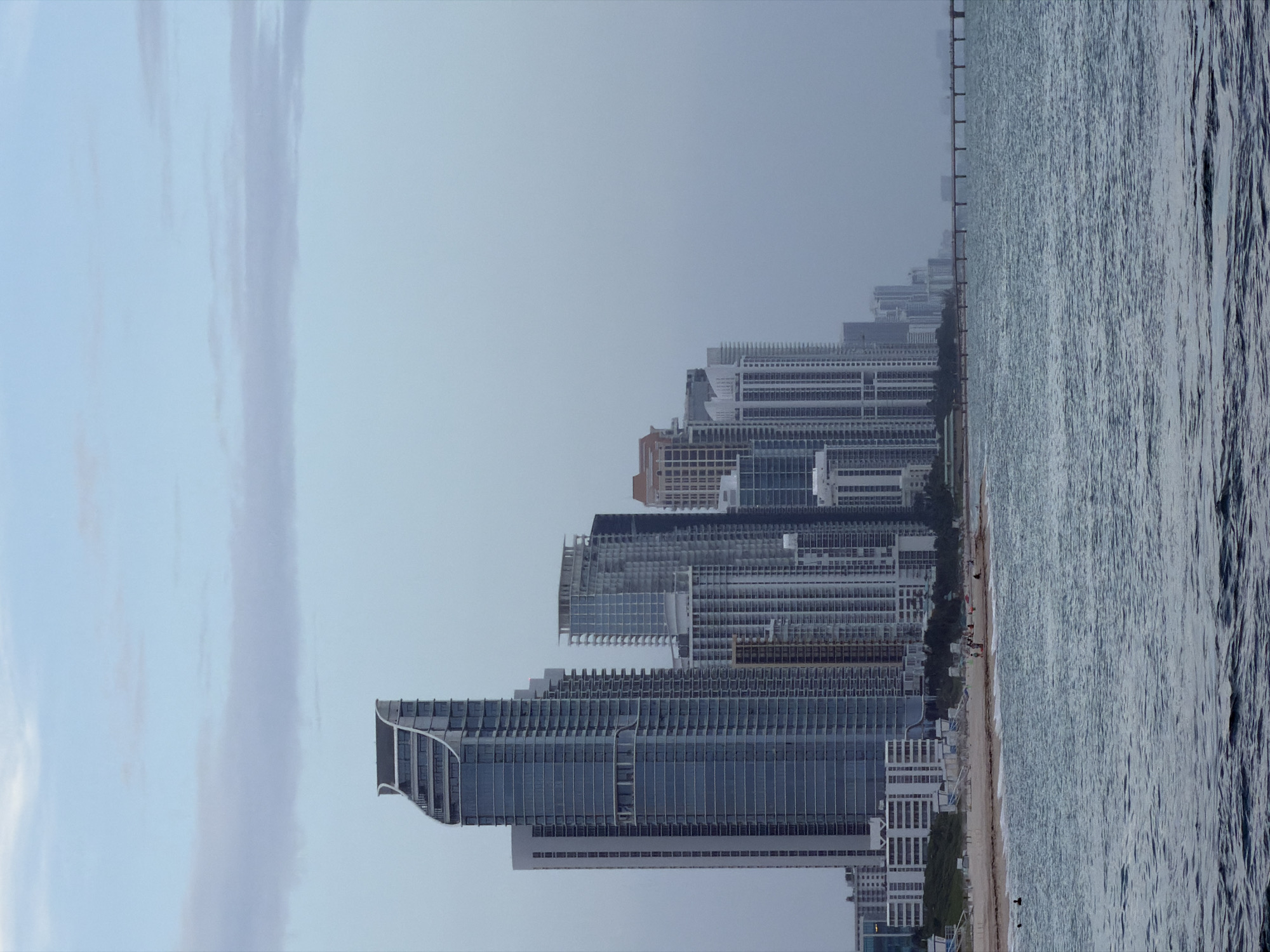This is a vertical, eye-level, medium-range photograph of a beachfront skyline in Bal Harbour Village, United States of America, on an overcast day. The ocean occupies the foreground and lower portion of the image, with gentle waves lapping at a sandy beach. The beach is lined with a row of modern, high-rise buildings of varying heights and architectural styles. The buildings are predominantly made of glass and concrete, with some featuring balconies and patterned facades. A long pier extends from the right side of the frame out into the ocean, with silhouetted figures visible on it. A few scattered individuals are visible on the beach, and one person is swimming in the ocean. The sky above is a pale blue, with soft, elongated clouds. The overall mood is tranquil and slightly muted due to the overcast conditions.