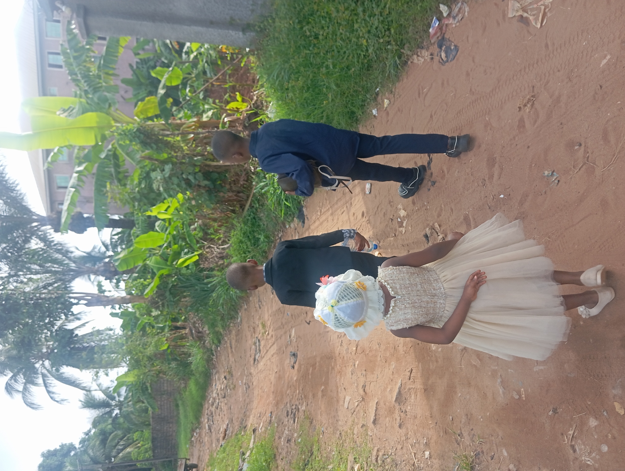 A ground-level shot shows three children walking on a dirt path in Nkpor, Nigeria. The path is surrounded by green vegetation, including banana trees. Litter is scattered on the path.
The first child, wearing a blue long-sleeved shirt and black pants, walks at the front. The second child, dressed in a black suit, holds a water bottle. The third child, wearing a cream-colored dress, a white hat, and white shoes, is positioned slightly behind and to the side of the second child.