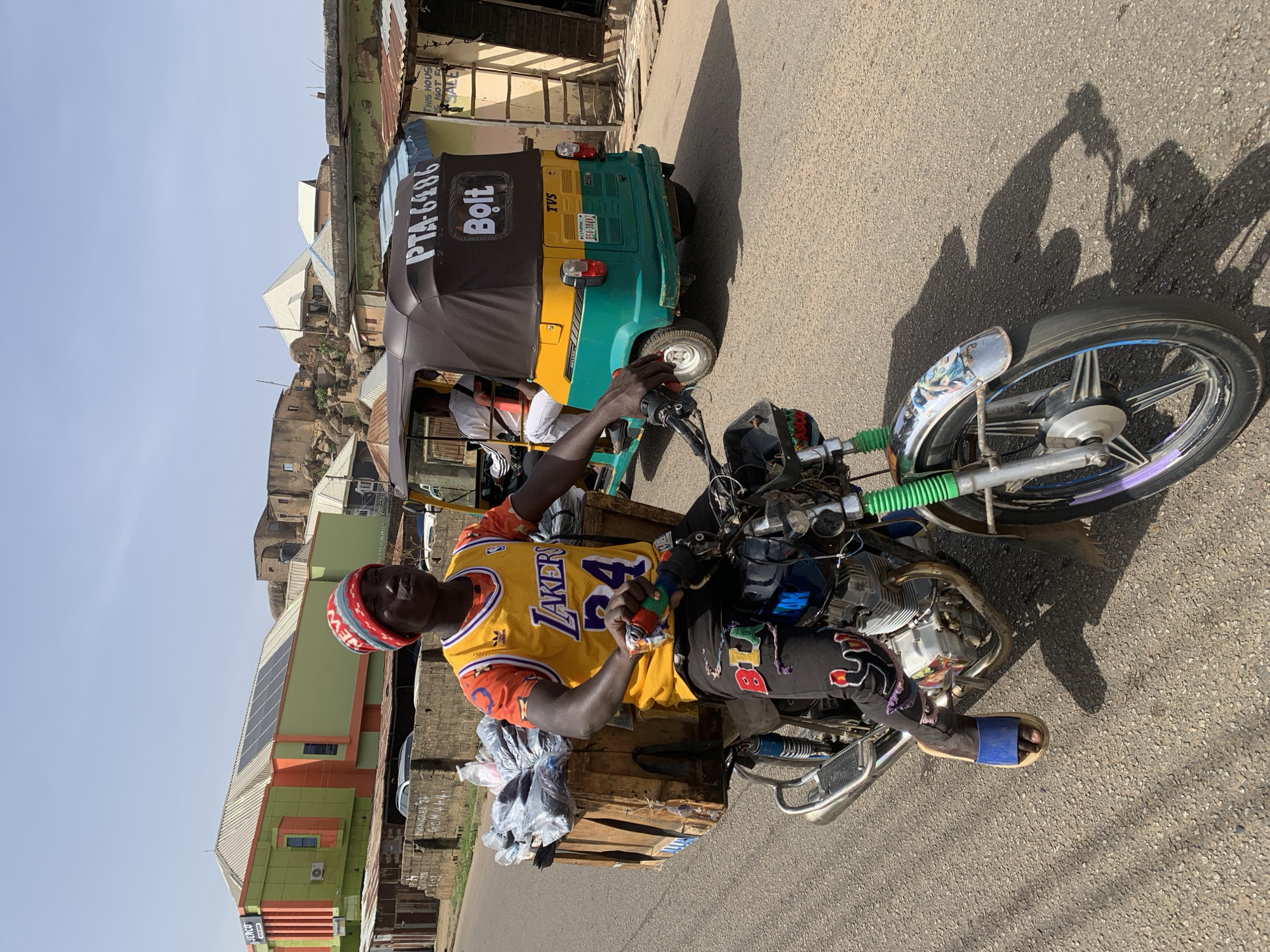 This image captures a bright, sunny street scene in Jos, Nigeria, focusing on transport and daily life. In the foreground, a man sits on a motorcycle, wearing a yellow Los Angeles Lakers jersey with "LAKERS" and the number "24" clearly visible, along with black pants and blue slide sandals. He also wears a red and white knitted hat. His motorcycle appears to be used for carrying goods, with a wooden crate and plastic bags attached to the rear. The rider holds an object with multiple colorful sections in his right hand. Strong shadows suggest it is midday or early afternoon.
Behind the motorcycle is a green and yellow motorized tricycle, commonly known as a Keke Napep. Its black canopy prominently displays the ride-hailing service "Bolt" and the identification number "PTA-6486." The rear of the tricycle shows "TVS" branding and a license plate with "RSA-38412" and the word "Jos," confirming the location context. A person is visible inside the Keke Napep.
The background features various buildings with different architectural styles, including structures with tin roofs and a more modern-looking building with solar panels on its roof and a sign for "JENCO." Rocky terrain and distant structures are visible under a clear blue sky. A faded sign on a building reads, "THIS HOUSE NOT FOR SALE." The road surface is asphalt, and there are additional markings and text, including "BLU" on the rider's pants.