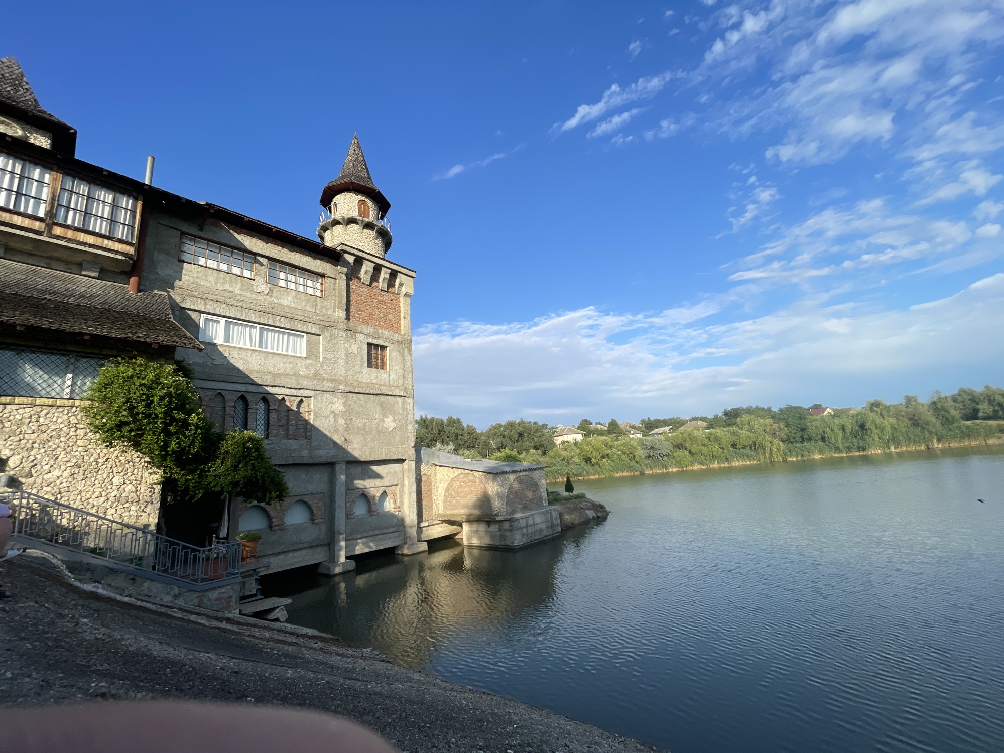 In the city of Colibași, Moldova, a photograph captures a building with castle-like architecture situated beside a body of water. The building features a prominent tower with a conical roof, multiple windows, and varying textures of stone and brick. A stairway leads up to an entrance on the left side of the building, and an area appears to be built into the water. The water's surface is rippled, reflecting the clear blue sky with scattered white clouds and the surrounding lush green trees and vegetation. Houses are visible in the distance along the opposite bank. The overall scene suggests a serene, somewhat rustic setting.