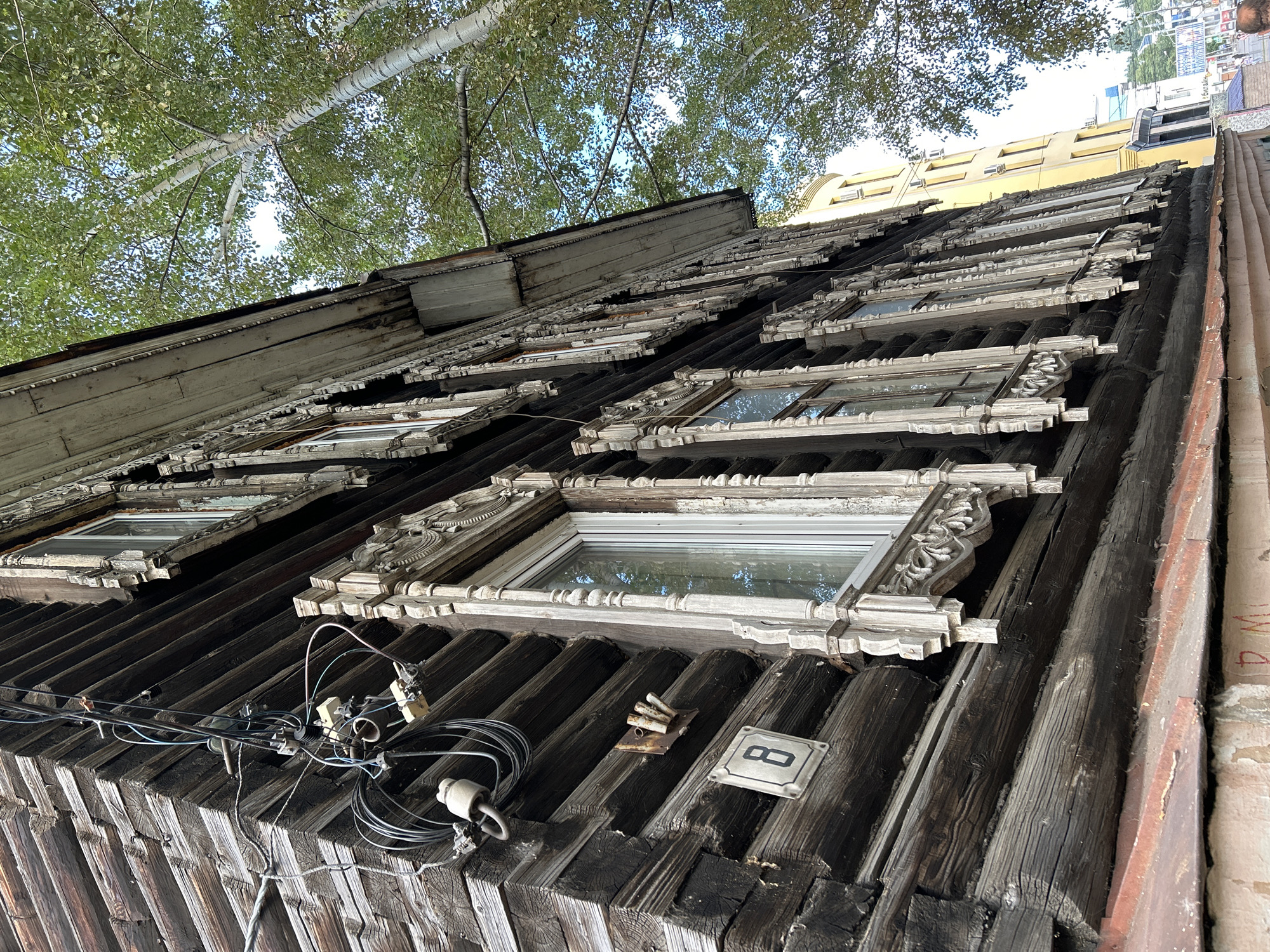 The image shows a close-up, low-angle view of an old, weathered wooden building with ornate window trim, located in Tomsk, Russia. The building's logs are dark and aged, with visible grain and texture. Several windows with intricate, light-colored carved wooden frames are visible along the side of the building.
In the upper left corner, a complex network of electrical wires and ceramic insulators is attached to the wooden wall. A square house number plaque with the numeral "8" is affixed to the logs below the wires.
In the background, a modern yellow building stands out against a bright sky, with the green foliage of a large tree partially obscuring it. The tree appears to be a birch, with its characteristic white bark. Some indistinct structures and possibly distant figures are visible further in the background, suggesting an urban setting. The overall impression is one of historical architecture juxtaposed with modern development.