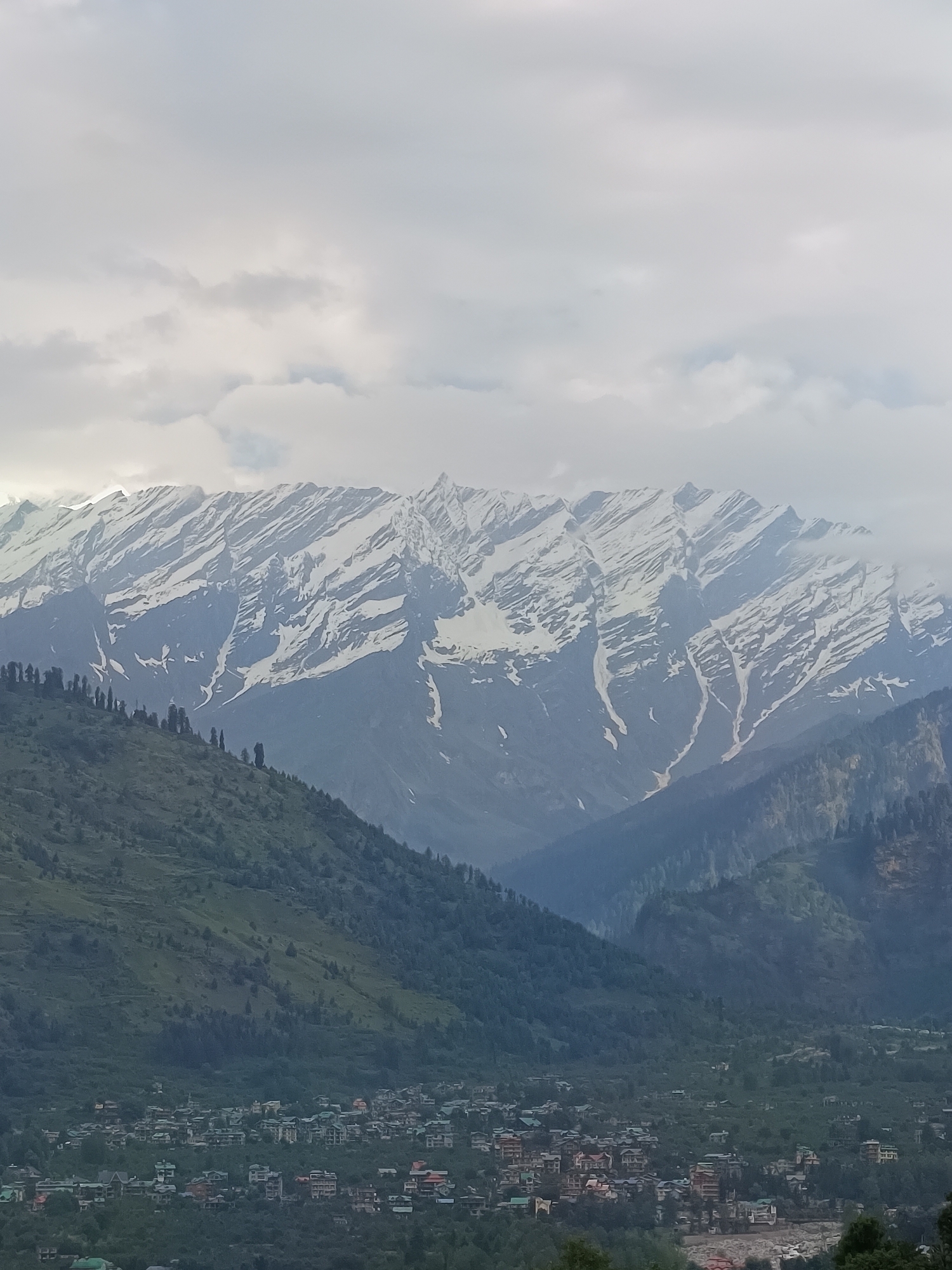 The image captures a sweeping view of a valley at the foot of snow-capped mountains. In the foreground, a densely populated town with multi-story buildings, many with reddish-brown roofs, is nestled amongst lush green hillsides. Tall, dark green coniferous trees dot the landscape, particularly on the slopes leading up to the higher elevations. The middle ground is dominated by the steep, verdant slopes of the valley, with the town receding into the distance. The background is defined by a massive mountain range, its peaks partially obscured by clouds. The mountains are characterized by dramatic striations of snow and rock, suggesting a rugged and imposing natural formation. The sky above is overcast with a soft, diffused light, contributing to a somewhat muted color palette of greens, blues, and whites. The scene appears to be a natural landscape juxtaposed with human settlement, likely taken from a vantage point on a hillside overlooking the valley and the town of Vashisht, India, given the characteristic architecture and the mountainous terrain. No people or specific actions are discernible in the image.