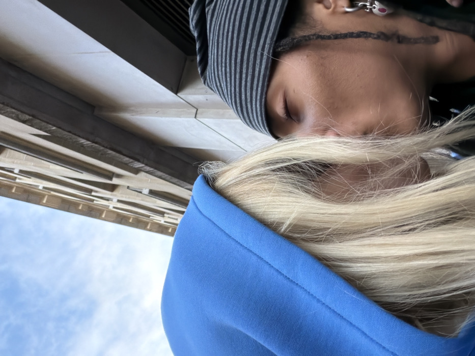 Two people are shown in a close-up shot, appearing to be standing outside. One person is wearing a striped beanie, with their eyes closed. A silver earring is visible. The other person, next to them, is wearing a bright blue hoodie. Their blonde hair is partially covering their face. In the background, a building is visible, along with a glimpse of the sky. The photo is taken from an unusual, tilted angle.