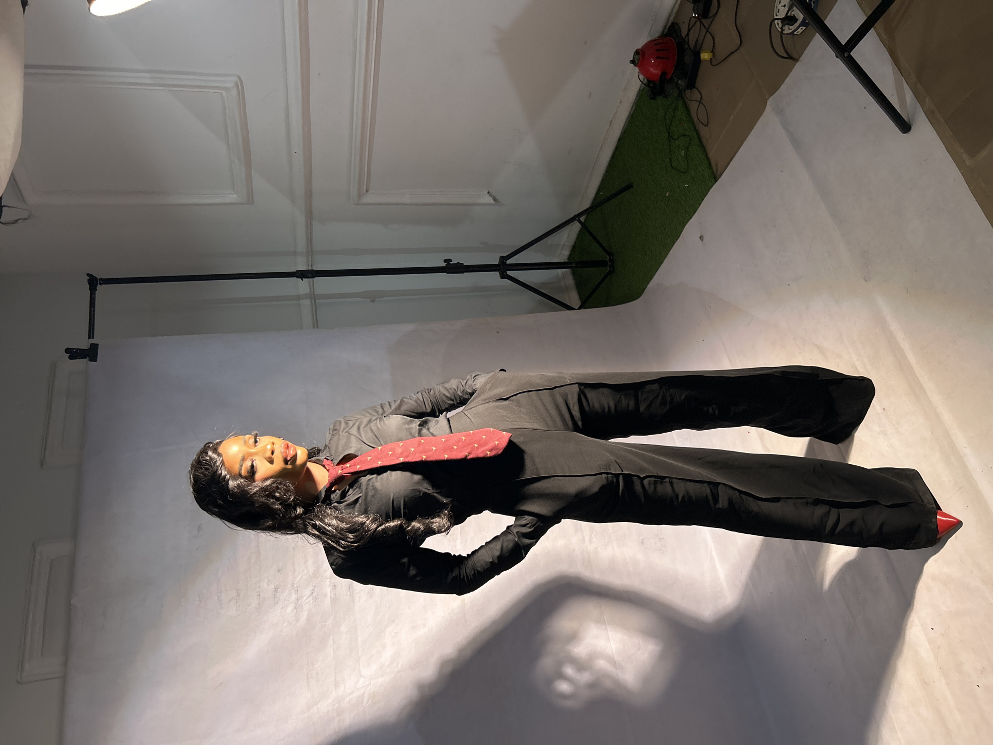 A woman is lying on her side on a white surface in a photo studio in Jos, Nigeria. She is wearing a black suit and a patterned red tie. The background consists of a white wall with decorative panels on the left, a green strip resembling artificial grass, and studio lighting equipment on the right. There is a black backdrop stand in the upper left. The lighting creates strong shadows on the white surface.