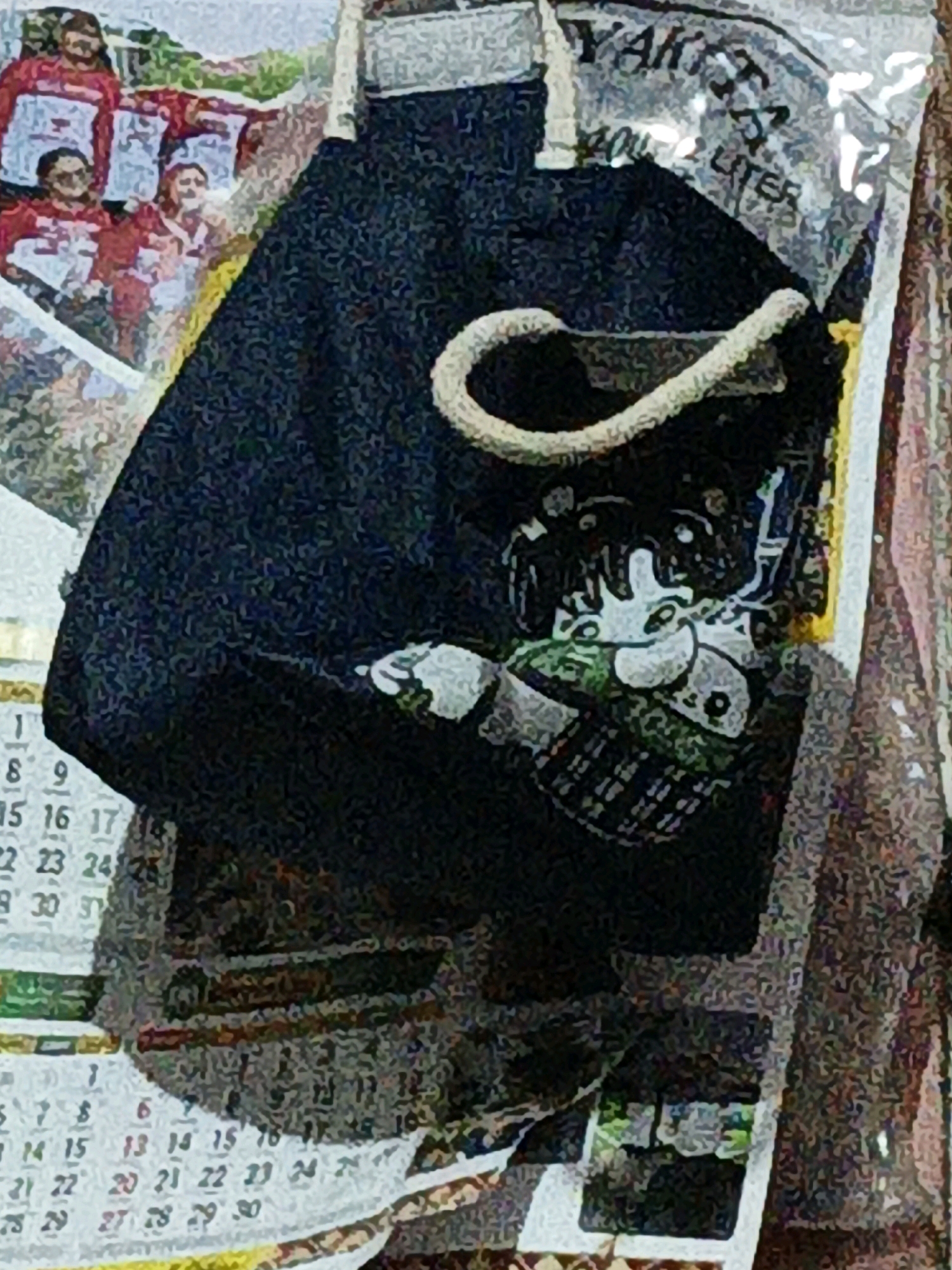 A dark fabric tote bag with a white rope handle is draped over a calendar in what appears to be a residential setting in West Jakarta, Indonesia. The bag has a cartoonish graphic printed on it. The calendar displays numbers and dates, suggesting a domestic environment. The overall lighting is dim, and the image quality is low, making detailed analysis difficult.