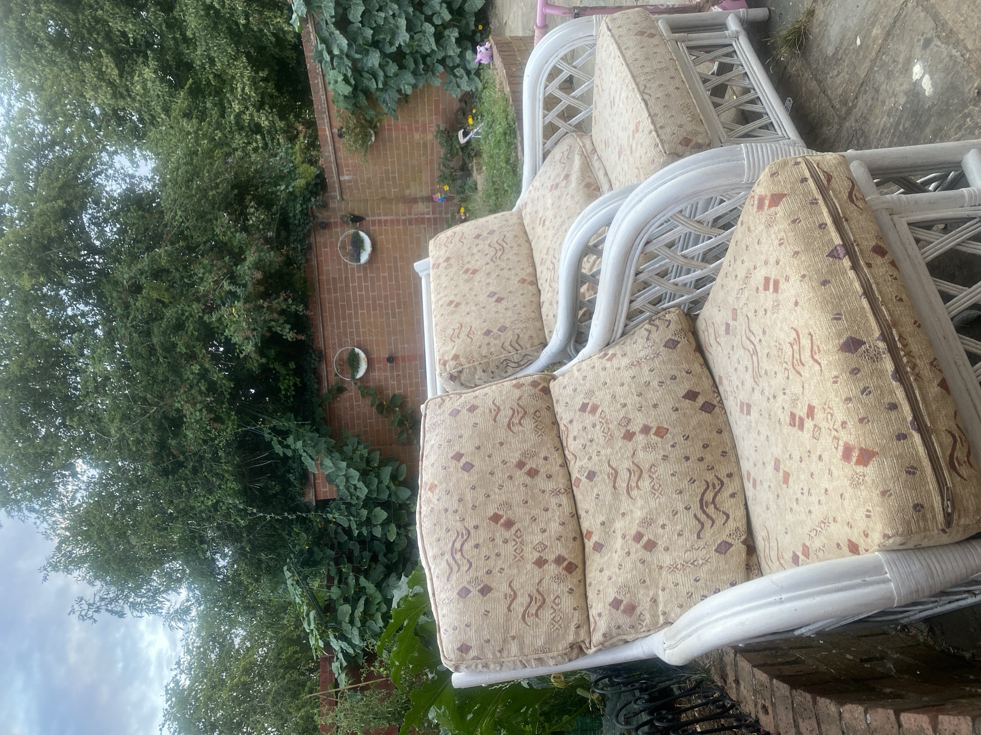 Two white wicker chairs with patterned cushions sit on a paved patio area. Behind the chairs is a brick wall with ivy growing on it and some planters. Trees and foliage form a backdrop, with a cloudy sky visible above. The overall scene depicts an outdoor seating area in a garden.