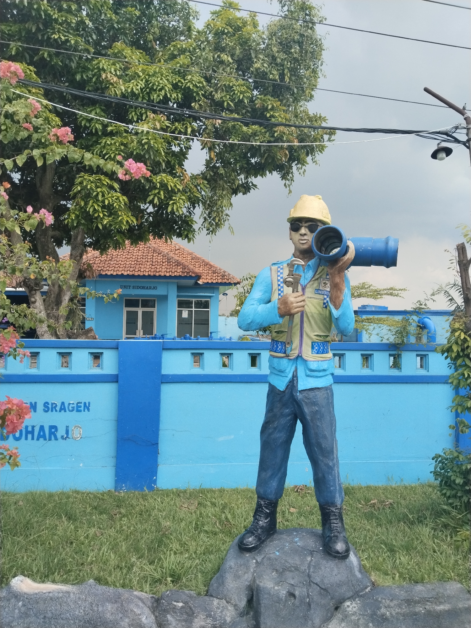 A statue of a construction worker, dressed in a light blue shirt and dark blue pants, is seen standing on top of a rock-like platform in Sidoharjo, Indonesia. He is wearing a yellow hard hat and sunglasses, and holding a blue pipe over his shoulder and tools in his other hand. Behind the statue is a blue building with the words "UNIT SIDOHARJO" inscribed above the doorway, surrounded by a blue wall with "EN SRAGEN BOHARJO" written on it. A tree with pink flowers is visible to the left of the building. Utility lines stretch across the sky above the scene.