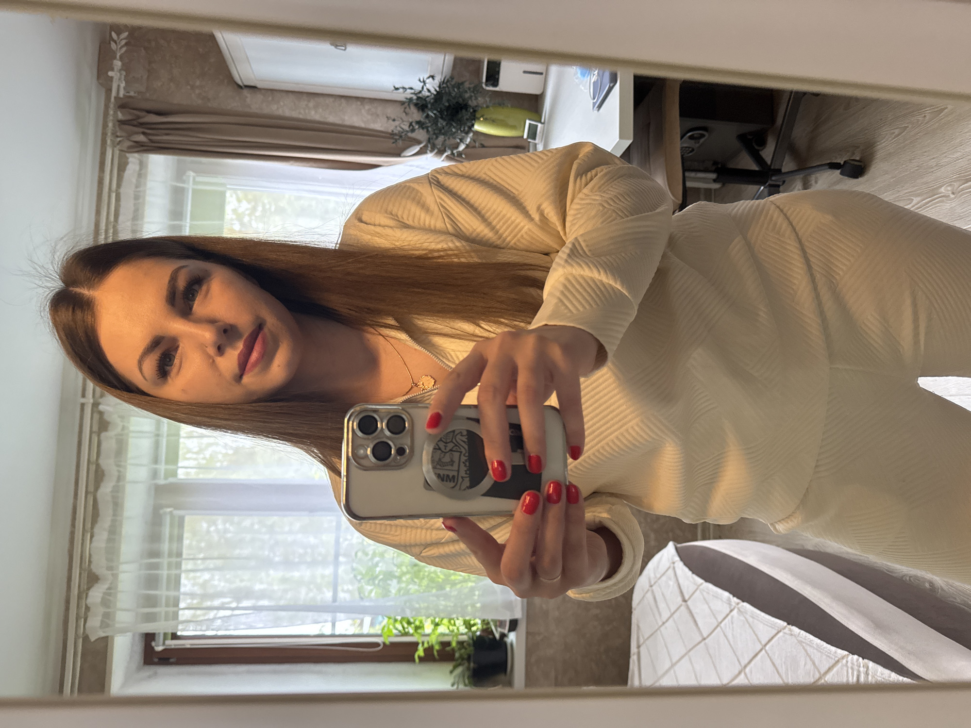 A woman is taking a selfie in a mirror. She has long brown hair and is wearing a cream-colored outfit. She is holding a phone with a white case and red nails. The background includes a window, a desk, and part of a bed. The location is Раздолье, Russia.