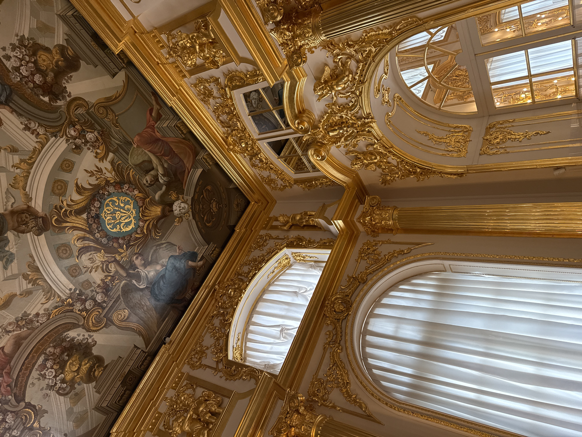 The image depicts the opulent interior of the Catherine Palace in Pushkin, Russia, specifically the White Hall. The scene is dominated by elaborate gold ornamentation covering the walls and ceiling. The ceiling features a fresco depicting angels and floral motifs, rendered in muted colors contrasting with the dominant gold.
The walls are adorned with gilded carvings, featuring intricate scrollwork, cherubs, and architectural details. Several large, arched windows are visible, draped with sheer white curtains that diffuse the natural light. These windows are framed by the same gilded ornamentation. In the upper right portion of the image, a smaller window with multiple panes reveals a reflection of the ornate ceiling and wall decorations.
The perspective is a low-angle shot looking upwards, emphasizing the grandeur and height of the hall. The overall impression is one of extreme luxury and baroque or rococo style. No people are visible in the image. The environment appears to be well-maintained and preserved, suggesting it is a historical site or museum.