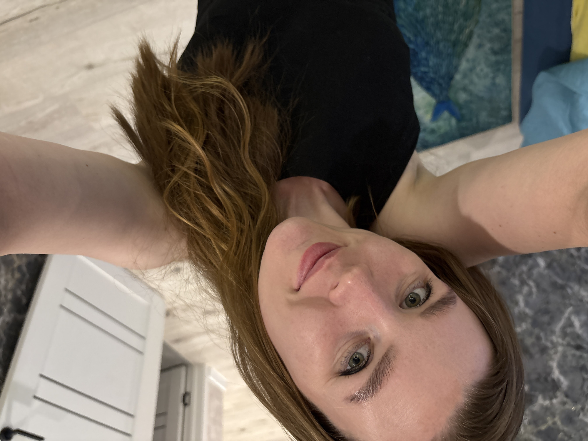 A fair-skinned woman with shoulder-length brown hair is posing for a selfie. She is wearing a black shirt and is indoors. A white door with a black handle is visible in the background, along with a multi-colored rug or painting. The lighting appears to be from overhead. The location is unknown and there is no evidence the image was taken in Izhevsk, Russia.