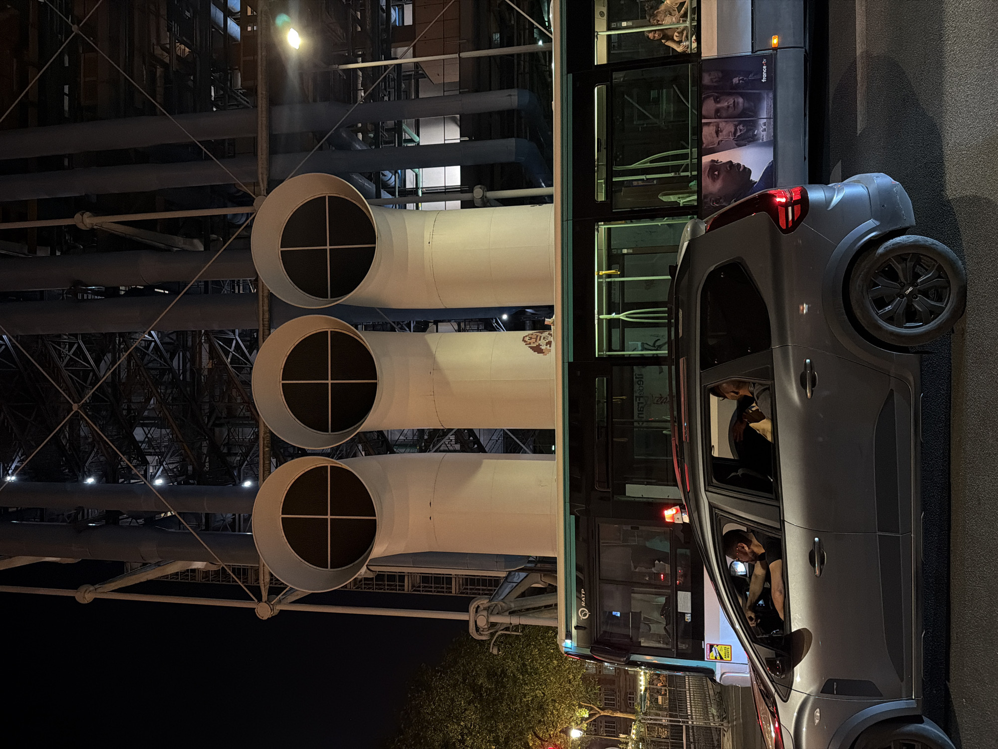 The image shows an exterior night scene in Paris, France, featuring the Centre Pompidou's distinctive exterior architecture with its exposed pipes and tubes. The perspective is looking up at the building. A city bus is positioned to the right of the frame, with an advertisement visible on its side. A gray car is parked or stopped beside the bus. Within the car, a person is visible in the driver's seat, possibly looking at a phone. Additional people might be present inside the bus. The light conditions are dark, illuminated by streetlights.