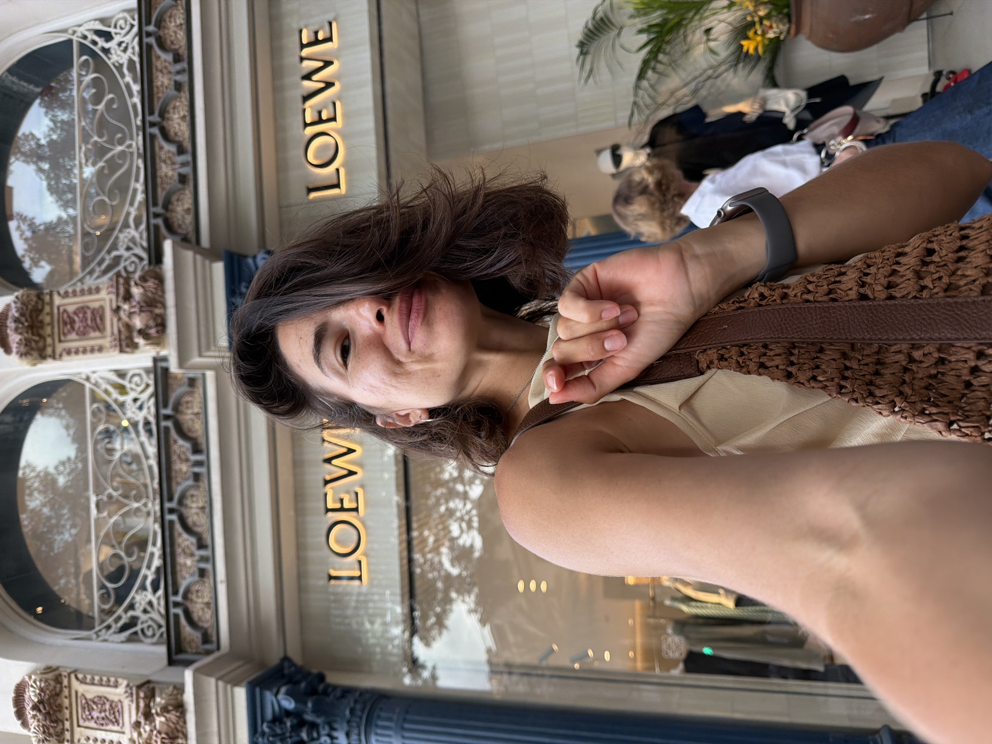 A woman is taking a selfie in front of a Loewe store. She has brown hair and is wearing a light-colored top. She is holding a woven brown bag and has a smartwatch on her left wrist. In the background, there are mannequins dressed in clothing. The store has a large window display and ornate architectural details above it. The overall setting appears to be an urban street.