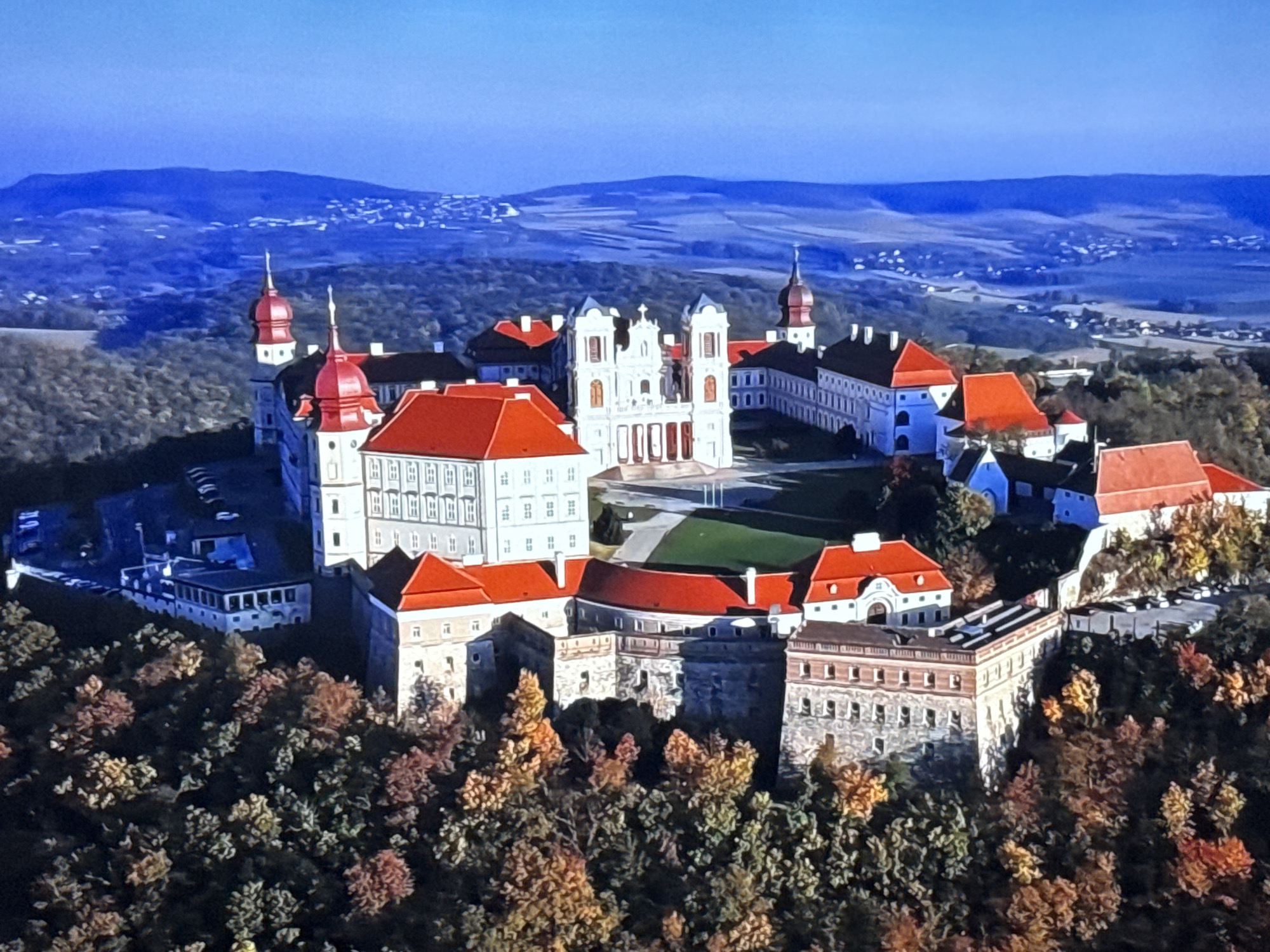 This is an aerial, high-angle, full shot of a large, sprawling, multi-building complex, likely a monastery or palace, situated atop a wooded hill with autumn foliage. The architecture features predominantly white walls with red tiled roofs, numerous windows, and several spires. The complex is surrounded by lush green trees displaying autumn colors of red, orange, and yellow. In the background, a vast, undulating landscape of rolling hills and distant settlements is visible under a clear blue sky. There are no people visible in the image. The location is not Burgas, Bulgaria, as the architectural style and landscape are not consistent with that region.
