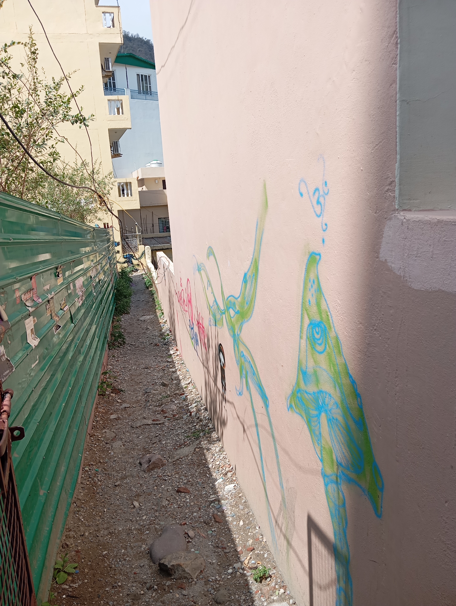 A narrow alleyway in Jonk, India, is depicted. The alley floor is composed of gravel and dirt. To the left is a green corrugated metal fence. To the right is a beige wall with graffiti in blue and green colors. Buildings are visible in the background beyond the alley.