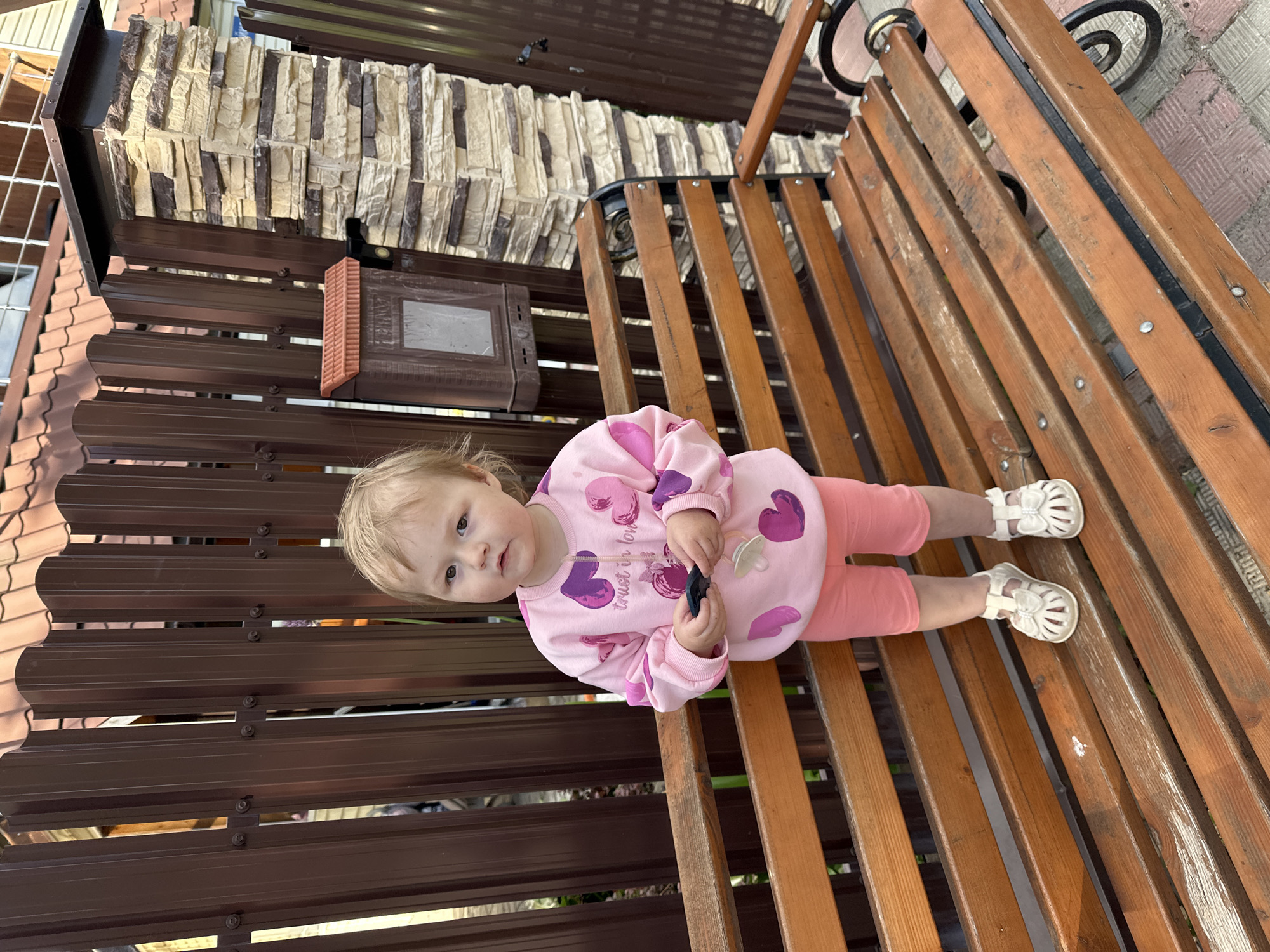 An outdoor scene in Nizhny Novgorod, Russia, features a young child, likely a toddler, sitting upright on a rustic wooden bench. The child has light-colored hair and is dressed in a pink long-sleeved top adorned with purple and pink heart-shaped patterns. The text "trust in love" is visible on the front of their top. They are wearing light orange leggings and white sandals with decorative bows. The child holds a small, dark-colored object, possibly a toy or remote control, in their hands, and a white pacifier is clipped to their shirt. Their expression is neutral and observant, looking directly at the viewer.
The bench, made of weathered light brown wooden slats, features black metal supports with decorative scrollwork. Behind the child and the bench is a dark brown corrugated metal fence or wall. Above this fence, a structure with textured, light-colored stone cladding, composed of rectangular blocks, is visible. A dark brown box-like object, possibly a small mailbox or speaker, is mounted on the fence structure. The ground visible next to the bench is paved with light-colored, patterned bricks. The scene is brightly lit, suggesting daytime.