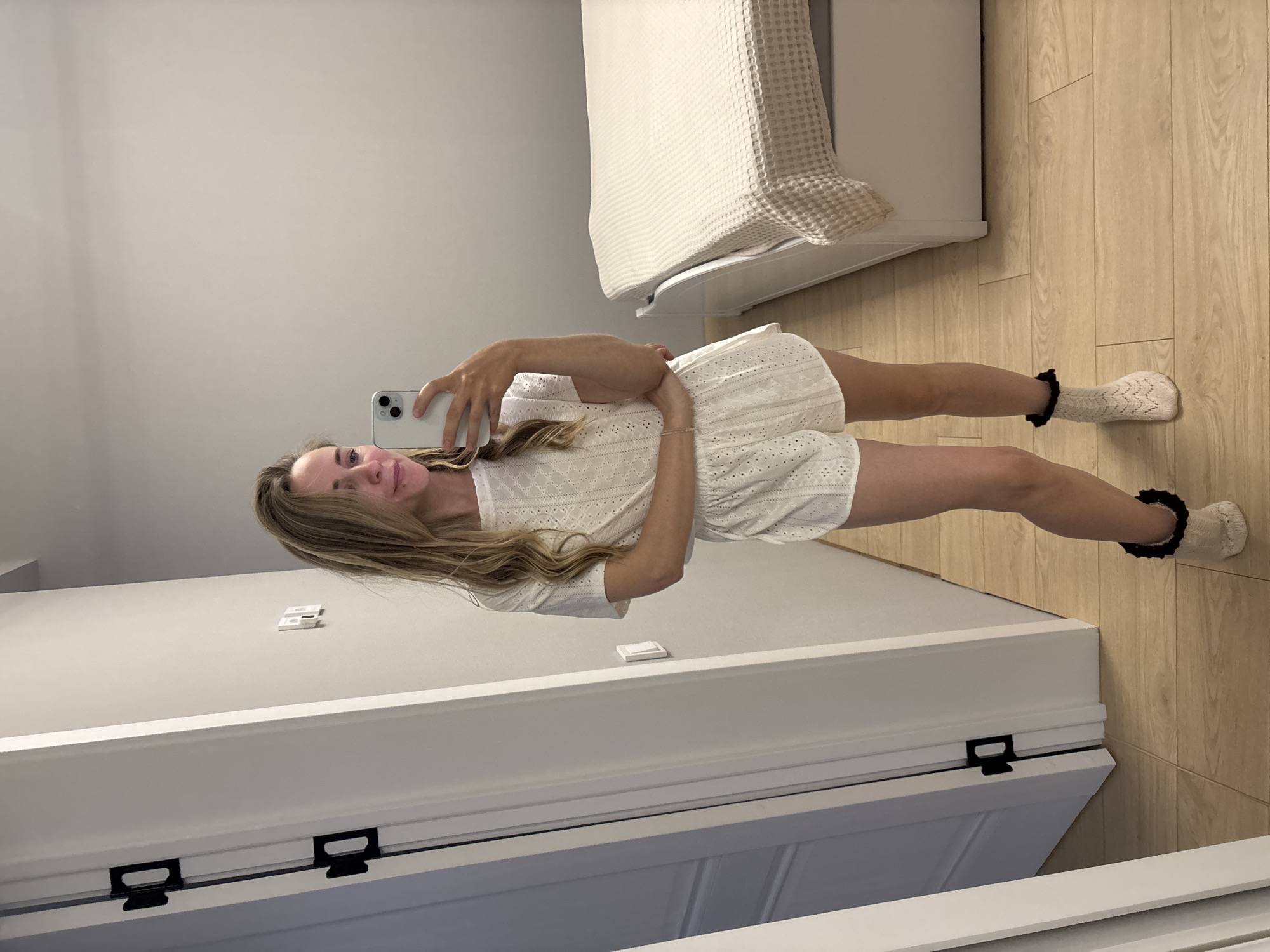A woman is seen taking a selfie indoors. She is wearing a white lace romper and white socks with black frills. The environment includes a bed with a white blanket, light-colored wooden flooring, and a white door with black handles. The location of the image is Chaikovsky, Russia.