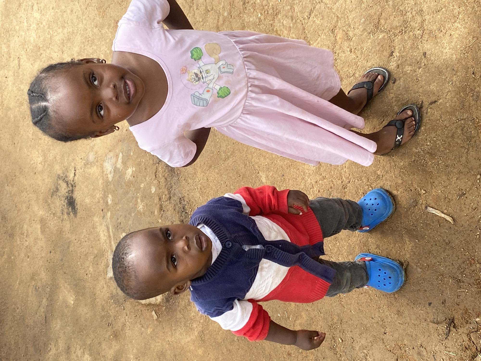 Two young children are lying on the ground. The older child, a girl, is wearing a light pink dress with an image of Sandy Cheeks from Spongebob Squarepants on it and black sandals. She is looking up at the camera and smiling. The younger child, a boy, is wearing a red, white, and navy cardigan, grey pants, and blue Crocs. He is also looking up at the camera. The ground is dirt. Given the ethnicity of the children, the location is likely Jos, Nigeria.