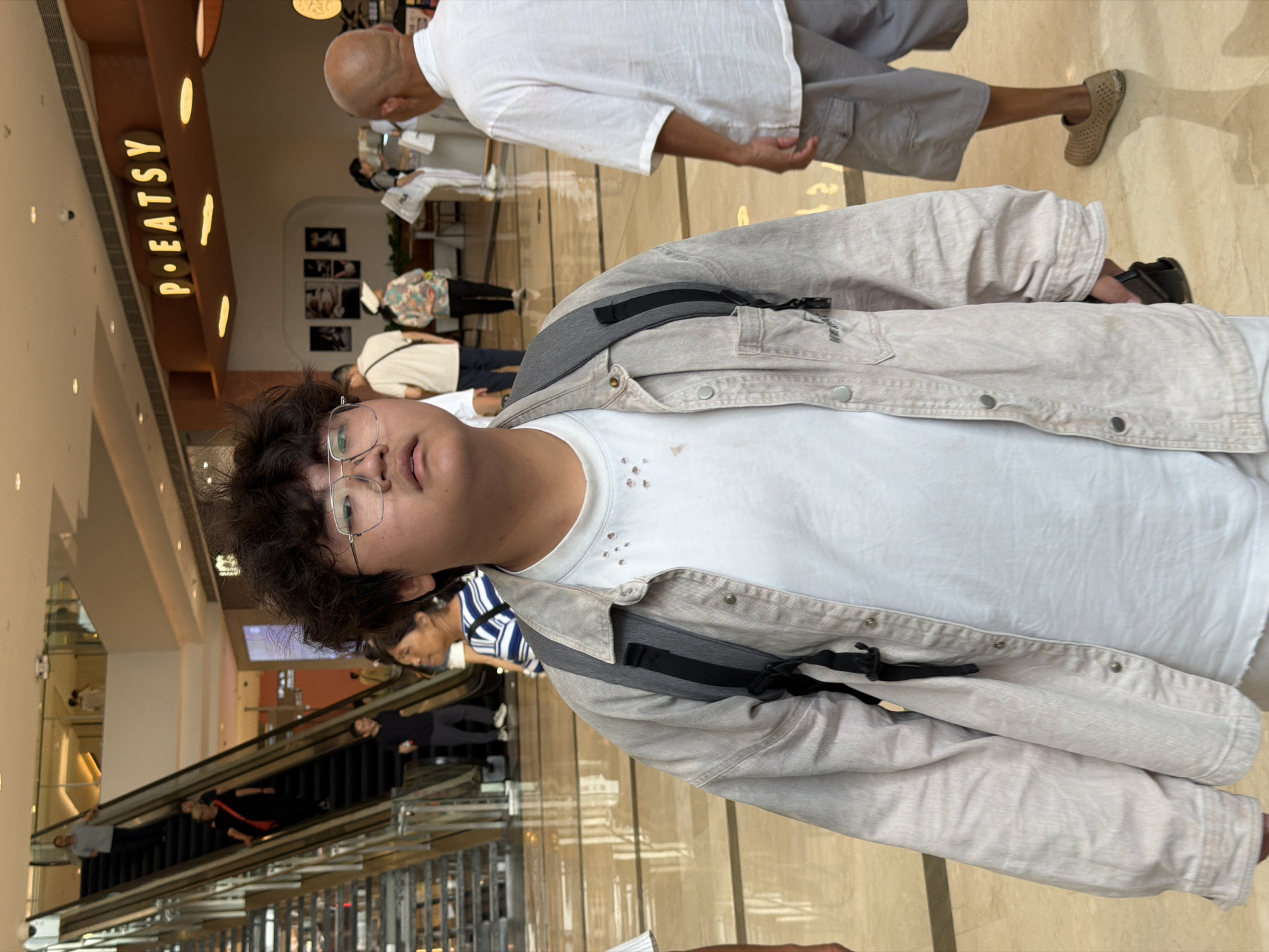 A young person with dark, curly hair and glasses stands in the foreground of a bright, modern indoor environment, likely a shopping mall in Yuzhong District, China. The person is wearing a light-colored, unbuttoned jacket over a white t-shirt with some light-colored markings around the collar, and a backpack with dark straps is visible. Their gaze is directed upwards and slightly to the right.
In the background, an escalator with people on it is visible to the left, with a man in a dark shirt and pants descending. To the right of the escalator, a woman wearing a striped blue and white shirt is partially visible. Further in the background, a sign with illuminated letters spelling "P•EATSY" is mounted above an establishment. Several other individuals are scattered throughout the area, some walking and others standing. The floor is made of reflective, light-colored tiles, and the overall lighting suggests it is daytime.