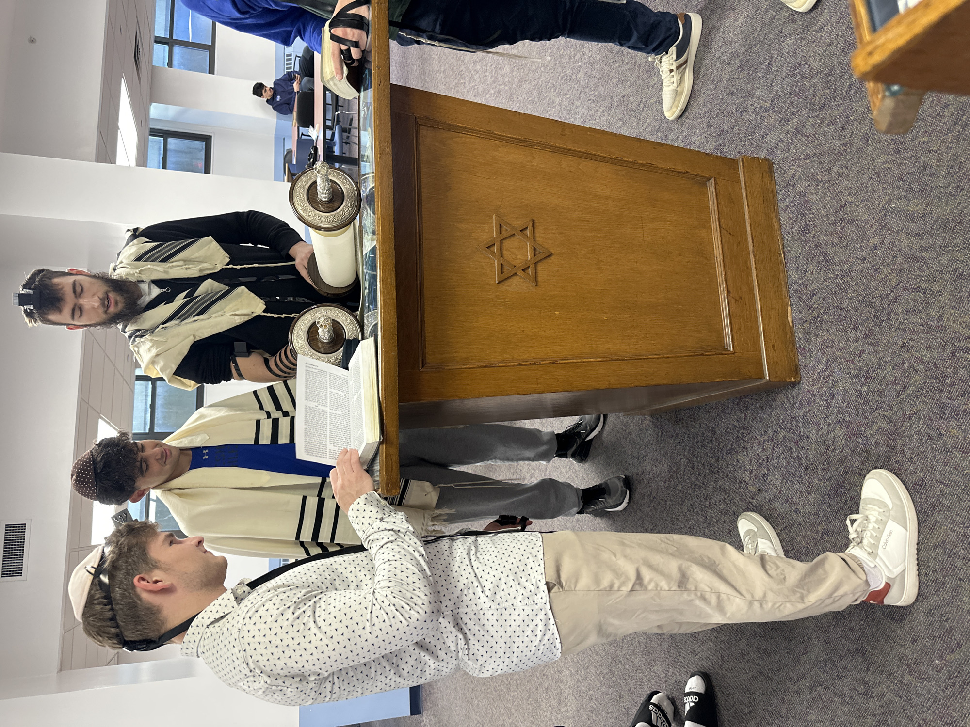 An indoor scene depicts four men engaged in what appears to be a Jewish prayer or study session. In the foreground, three men are visible around a large, ornate wooden lectern or stand featuring a carved Star of David on its side.
To the left, a man with a beard and a black zip-up top wears a Tallit (prayer shawl) draped over his shoulders and a Tefillin (phylactery) box strapped to his forehead. He looks intently towards an open Torah scroll that rests on the stand, which has decorative silver covers on its handles.
To the right, another man, wearing a white patterned button-down shirt, khaki pants, and a white kippah (skullcap), holds an open book, likely a prayer book or religious text. He is looking towards the Torah and the bearded man. He wears white sneakers with "Calvin Klein" visible on one shoe. Just to his left, partially obscured, a third man wearing a blue shirt (with an "Under Armour" logo visible) under a Tallit and a patterned kippah, also looks down at an open book.
The floor is covered with a grey carpet. In the background, another individual can be seen seated, and large windows are visible, suggesting a well-lit room. Adidas slides are also partially visible on the floor near the foreground. The overall atmosphere suggests a focused and reverent gathering for religious observance or learning.