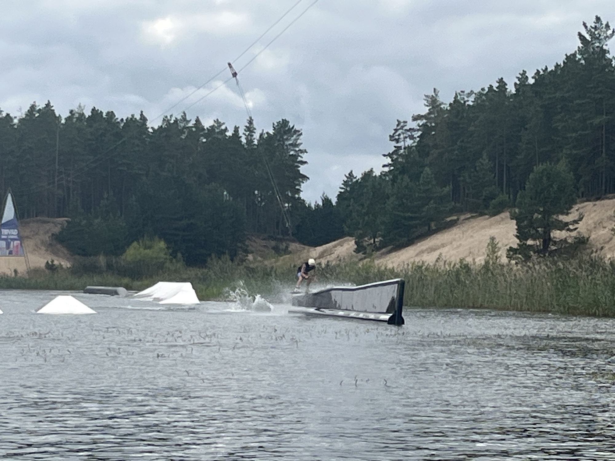 This image captures a wakeboarding scene at the Männiku wake park in Männiku, Estonia. A wakeboarder, wearing a helmet and dark shorts and shirt, is in action, mid-air or transitioning over a large, angled, dark-colored ramp. Water is spraying up around the ramp and the wakeboarder, indicating movement and activity. In the foreground, the surface of a body of water, likely a lake or pond, is visible with ripples and reflections. To the left of the wakeboarder, two white, pyramid-shaped structures are partially submerged in the water, serving as obstacles or starting points. Further in the background, a dense forest of pine trees lines the shore, with sandy hills visible beyond the trees. A cable system, typical of a wake park, extends across the sky, with a pulley system and a red component visible high up. A flag with blue and white colors and some text is also visible on the far left side of the image. The sky is overcast with scattered clouds.