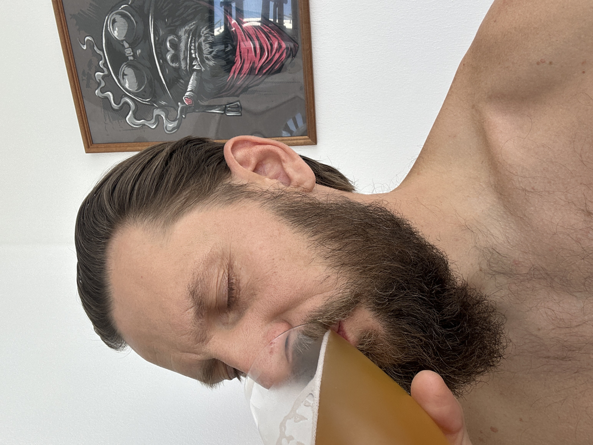 A man with a beard is drinking from a glass of beer. He is shirtless and his left shoulder and upper chest are visible. He has brown hair and closed eyes. In the background, there is a framed picture of a gorilla wearing a bandana and smoking a cigar. The image is likely taken indoors, possibly in a home, given the presence of a framed picture on a wall. The location is Vsevolozhsk, Russia.