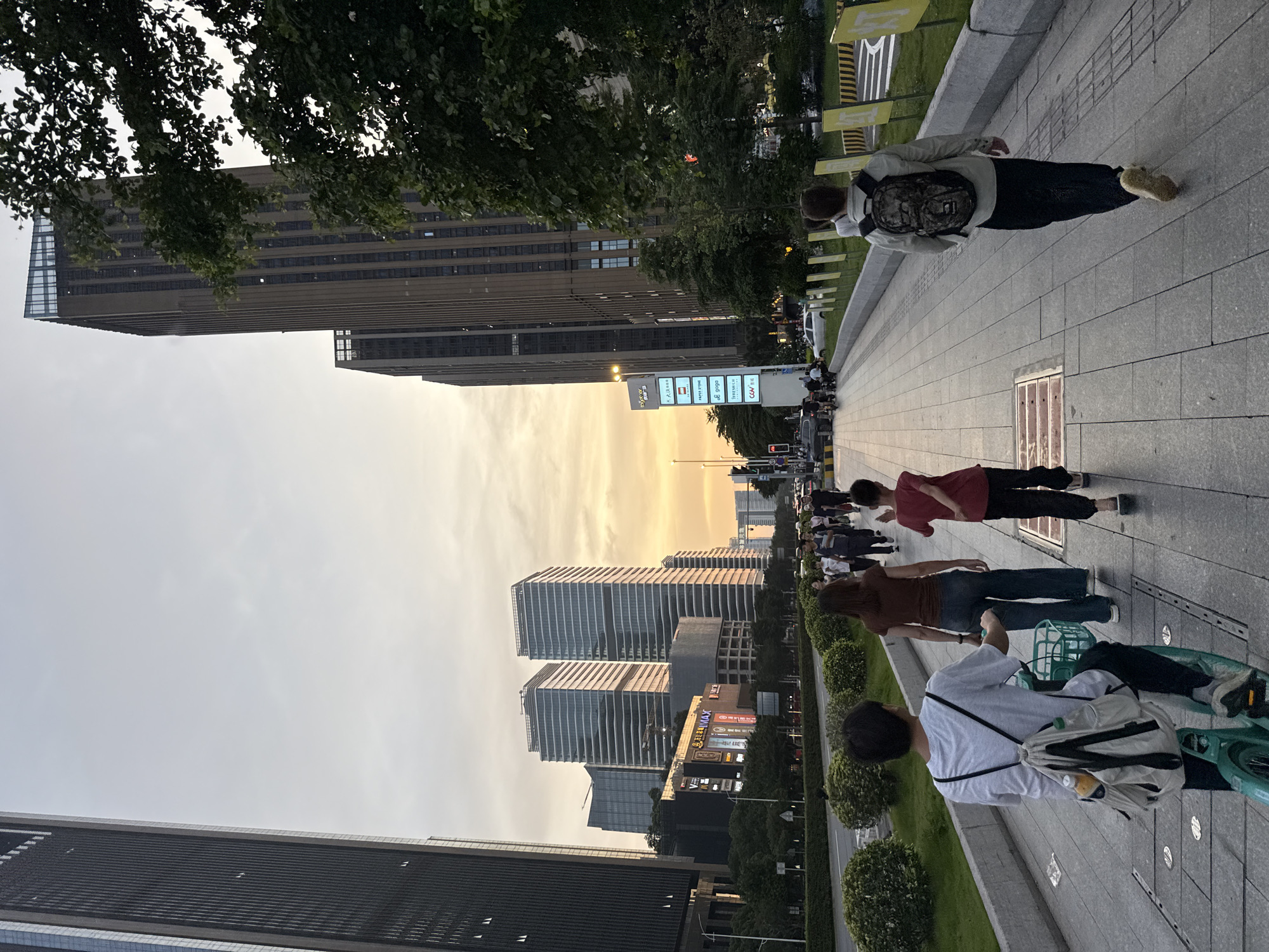 The image depicts a street scene in Haizhu District, China, at dusk or dawn. The sky is a pale gradient, with hints of orange. A group of people is walking along a wide, paved walkway next to a road with trees and grass. Modern high-rise buildings line both sides of the street. Some people are walking, while others are on bicycles. One person has a backpack on, and another has a basket on their bicycle. There's street signage and some greenery visible along the side of the walkway. The scene appears to be in a commercial or business district.