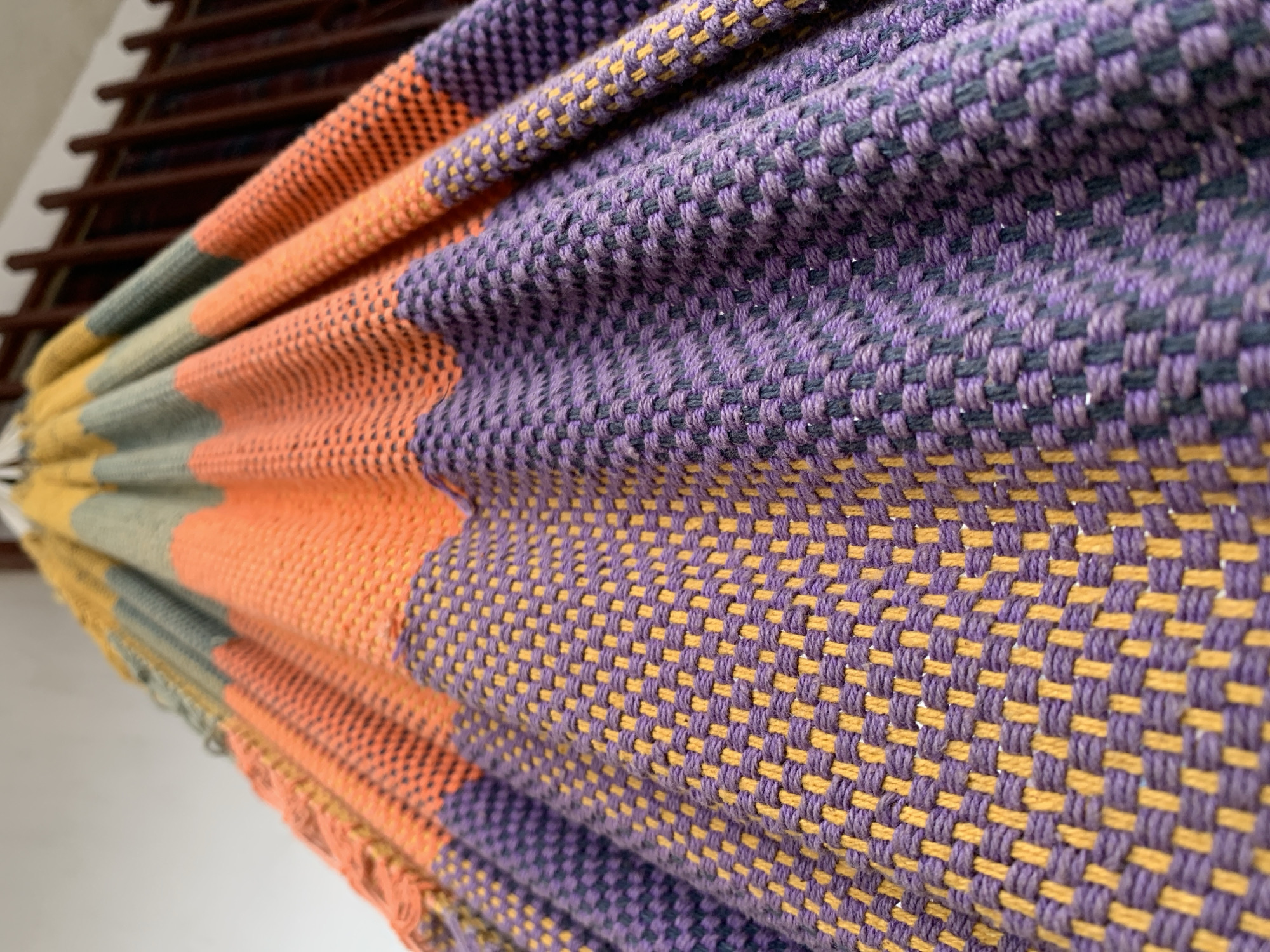 A close-up photograph captures a section of a vibrantly colored, intricately woven textile, likely a hammock, seen folded or draped. The fabric features distinct horizontal bands of color, including rich purple, bright orange, mustard yellow, and a muted olive green or grey. The prominent weaving technique creates a distinctive checkerboard pattern.
Detailed examination reveals specific color combinations within the weave; for example, some purple sections are tightly interlaced with mustard yellow threads, while other purple areas are woven with a darker, possibly black or dark grey, yarn. The orange bands also exhibit a similar detailed, contrasting weave. The texture of the fabric appears coarse and tactile.
In the background, a dark brown wooden structure, possibly part of a hammock stand, furniture, or a railing, is partially visible and out of focus. This wooden element is set against a bright, light-colored background, suggesting an indoor or shaded outdoor environment. The lighting is bright and even, indicating that the photo was taken during the day. There is no visible text in the image. Given the location context of Santa Ana, Venezuela, this colorful and robustly woven item is consistent with traditional hammocks from the region.