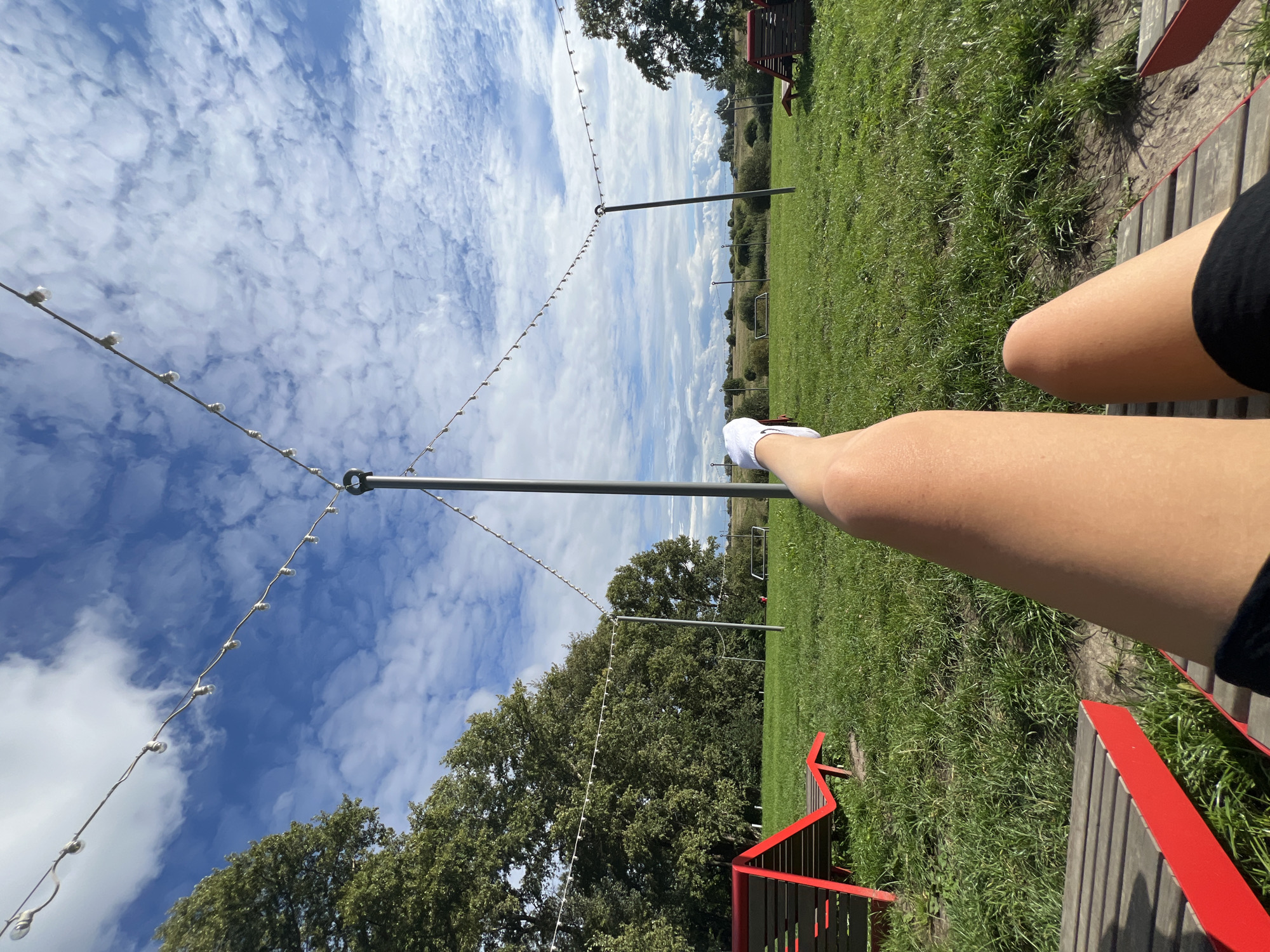 A first-person perspective shot shows a person's legs and feet extending towards a blue sky with scattered white clouds. The person is seated on a red and wood lounge chair outdoors. In the background, a grassy field is dotted with more lounge chairs, trees, and poles with string lights. The scene appears to be in a park or outdoor recreational area.