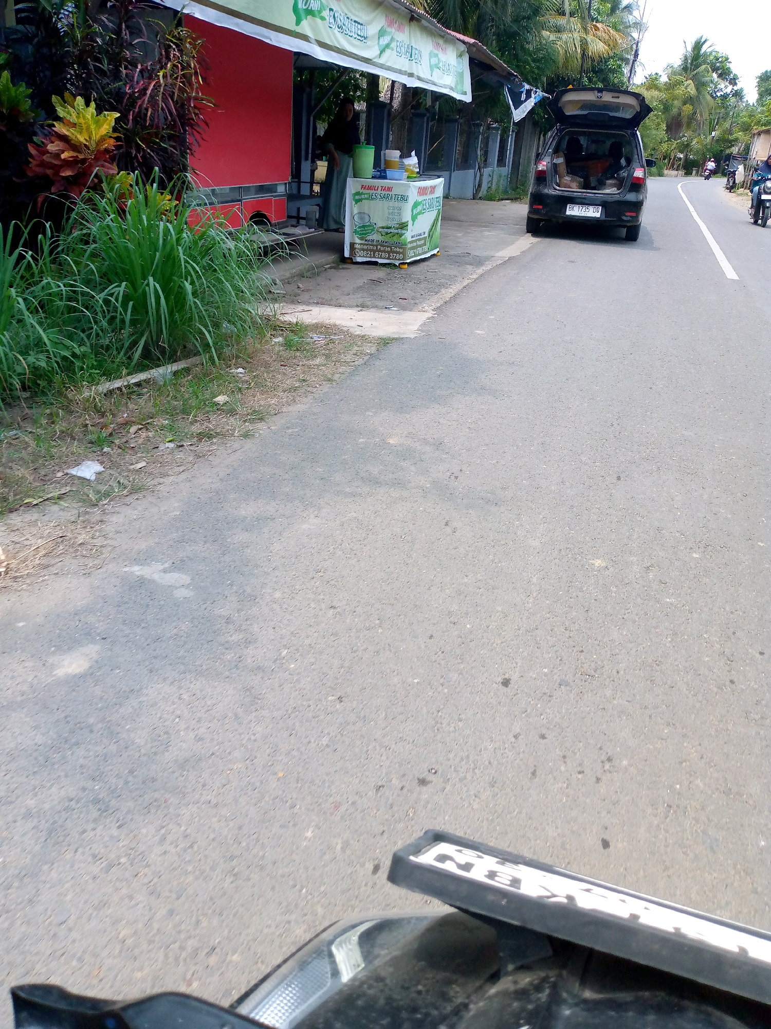 The image depicts a roadside scene in Blangkabu, Indonesia. In the foreground, the edge of a motorcycle is visible, with a partially obscured license plate reading "BN 30". To the left, a roadside stall with a red facade and a green and white banner is set up. The banner displays text including "FAMILI TANI" and "ES SARI TEBU". A woman in a dark headscarf and light-colored skirt stands behind the stall, tending to items on display. Lush green grass and plants grow alongside the road. Further down the road, a black car with its trunk open is parked. The car's license plate is visible as "BK 1735 DB". Two motorcycles are also present on the road, one ridden by a person in a blue shirt, and another, further away, also occupied by a rider. The road itself is paved and appears to be a two-lane street, with a white line marking the center. The environment suggests a sunny day with palm trees and other vegetation lining the street.