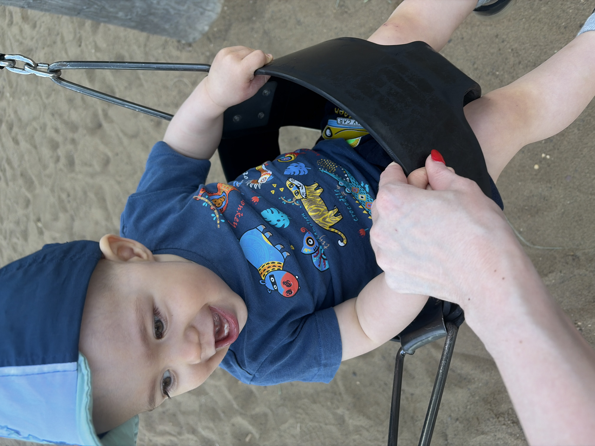 A baby wearing a blue t-shirt with animal prints and a blue hat is sitting in a black swing at a playground. A person's hands are visible, stabilizing the swing. The ground appears to be covered in sand. The location is Chaikovsky, Russia.