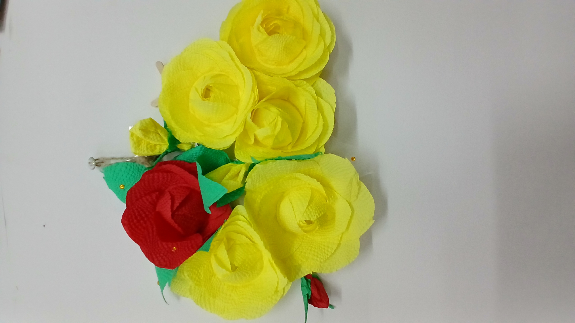 The image displays a handmade bouquet of flowers, predominantly yellow roses with a single red rose at the center. The flowers appear to be crafted from tissue or crepe paper. Green paper leaves are interspersed among the blossoms, and small gold beads are used as accents. The bouquet is attached to a white wall with a small silver tack. There is no indication of the city of Oral, Kazakhstan, or any specific location within it. The scene is static and focused solely on the floral arrangement.
