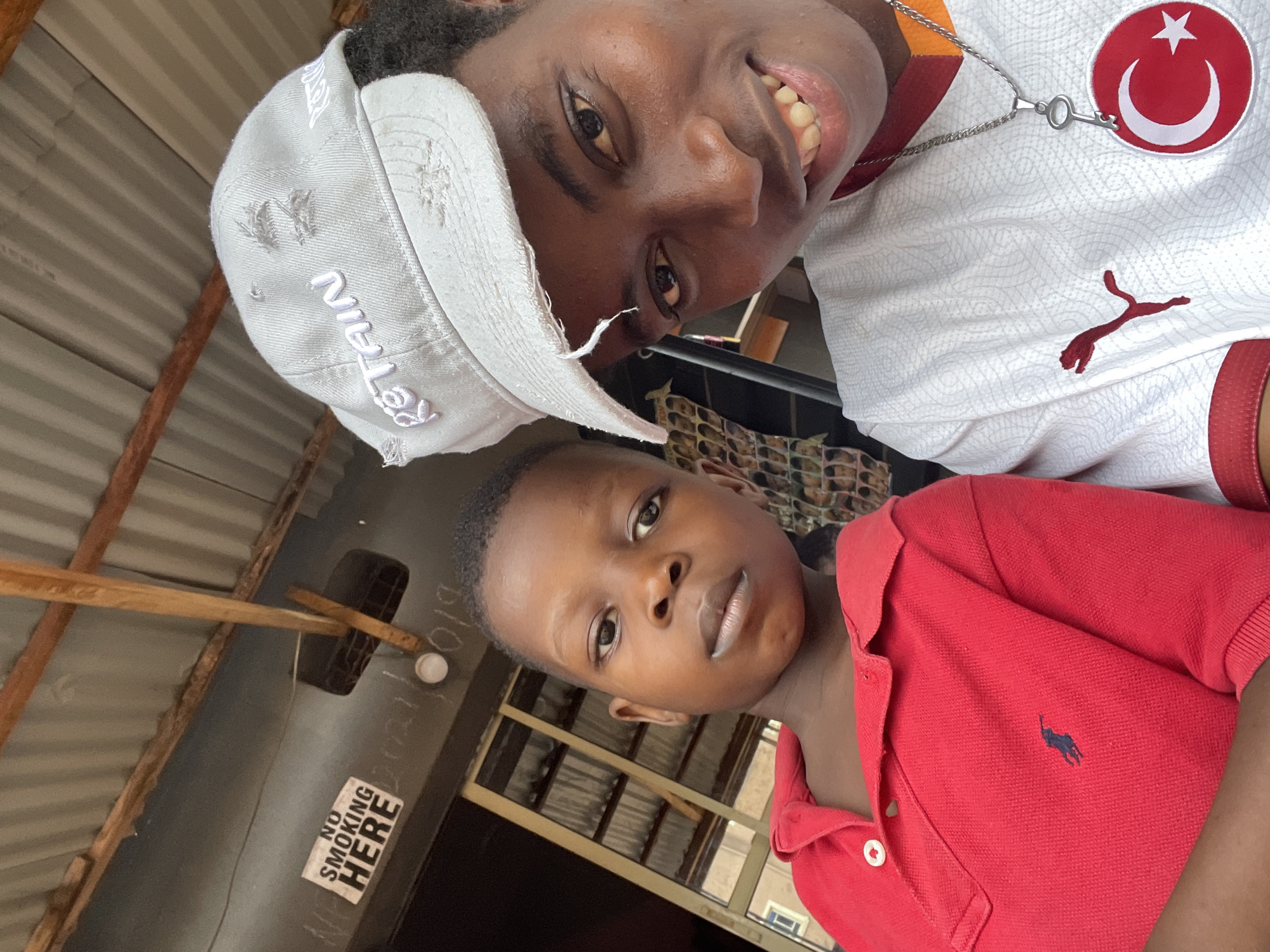 Two individuals are photographed in a close-up selfie setting. The person at the top of the frame wears a light grey, distressed baseball cap with the word "Retain" visible on the front. They also wear a white jersey with maroon accents and a Turkish flag pendant. This person is smiling. Below them is a younger person wearing a red polo shirt.
In the background, parts of a building are visible including a "No Smoking Here" sign. Based on the architectural style, the image was likely taken in Onitsha, Nigeria.
